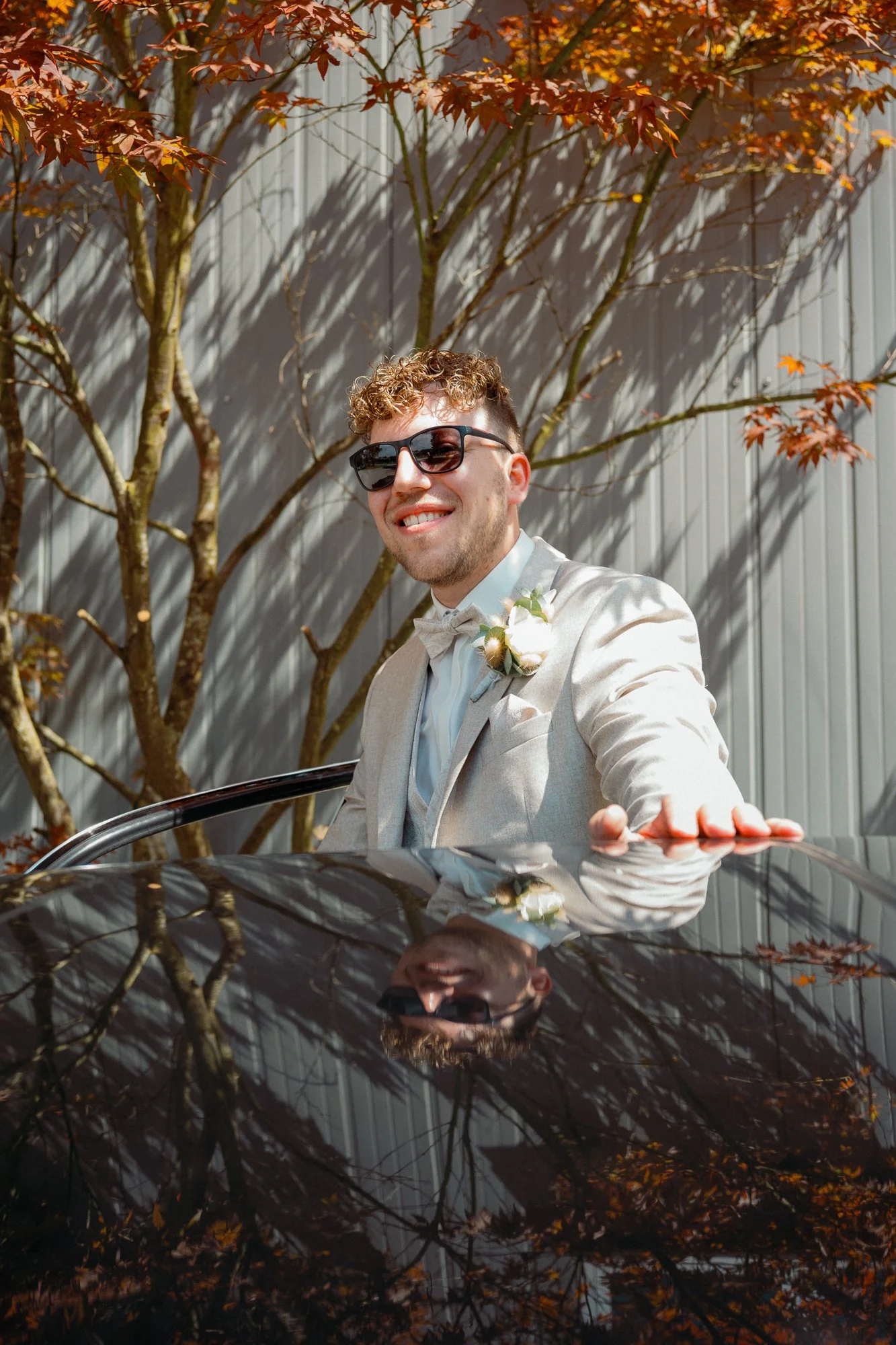 Junger Mann mit Locken, Sonnenbrille, weißem Anzug und Blume im Revers, posiert vor einer roten Ahornbaum und lacht, während er sich in einem schwarzen Auto mit reflektiertem Dach befindet.