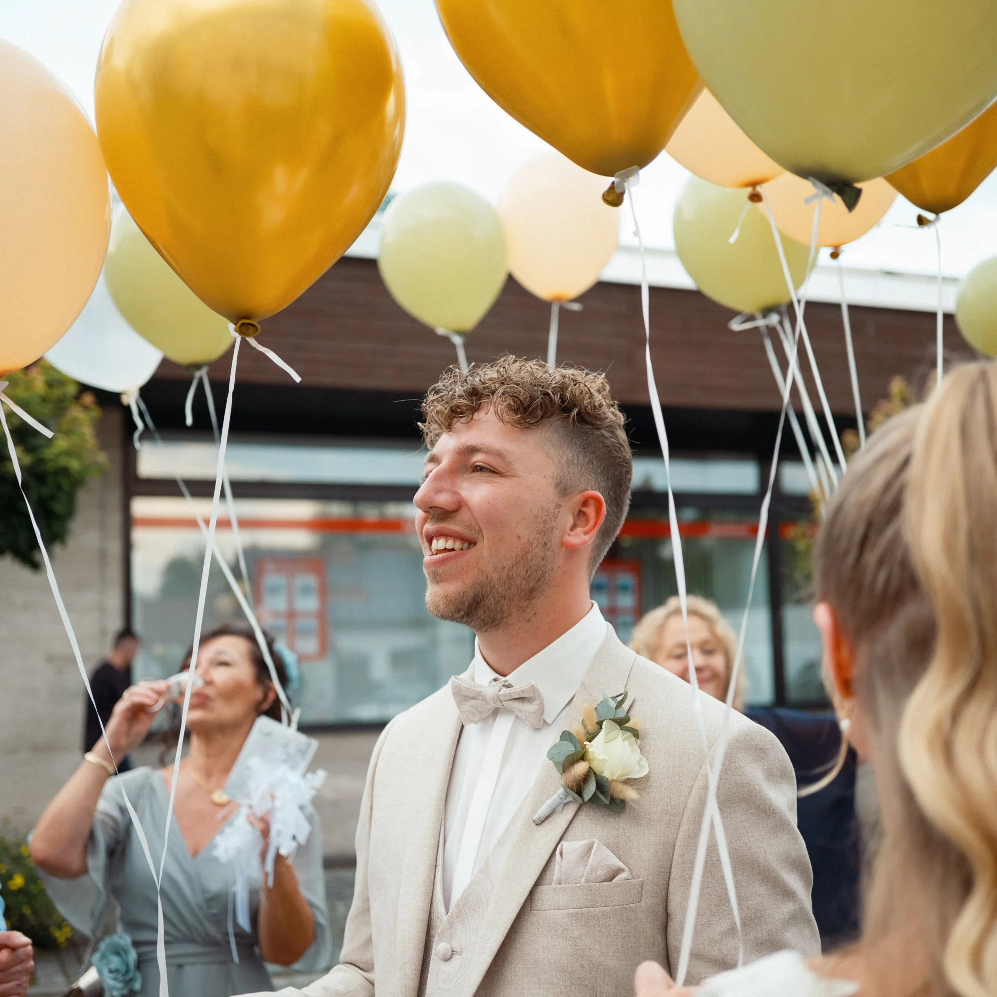 Ein junger Mann in einem weißen Anzug mit Fliege und Blumenschmuck im Revers, umgeben von bunten Luftballons, bei einer Feier im Freien.