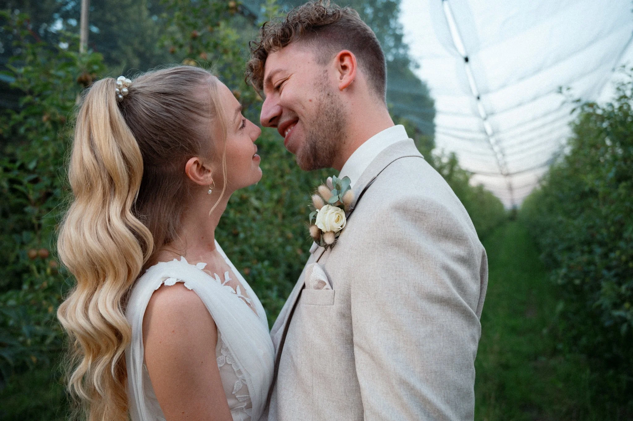 Ein Paar bei einer Hochzeit, lächelnd und sich gegenüberstehend, im Freien, umgeben von Bäumen.
