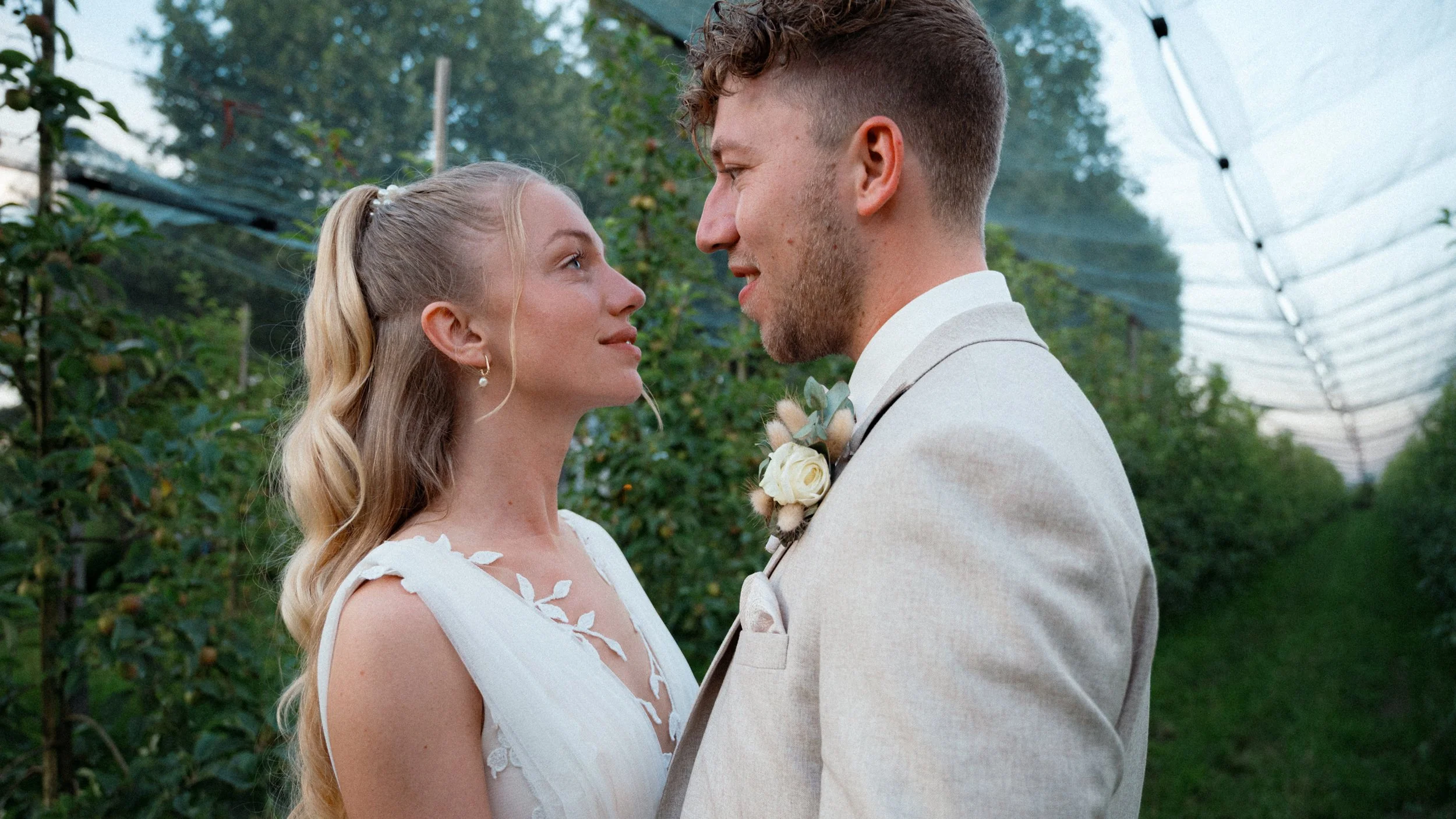 Ein junges Brautpaar steht sich gegenüber in einem Garten, blickt sich liebevoll an, die Frau trägt ein weißes Hochzeitskleid mit floralen Applikationen, der Mann trägt einen hellen Anzug mit Blumenbouquet. Im Hintergrund sind grüne Bäume und ein lichtdurchlässiges Zelt sichtbar.