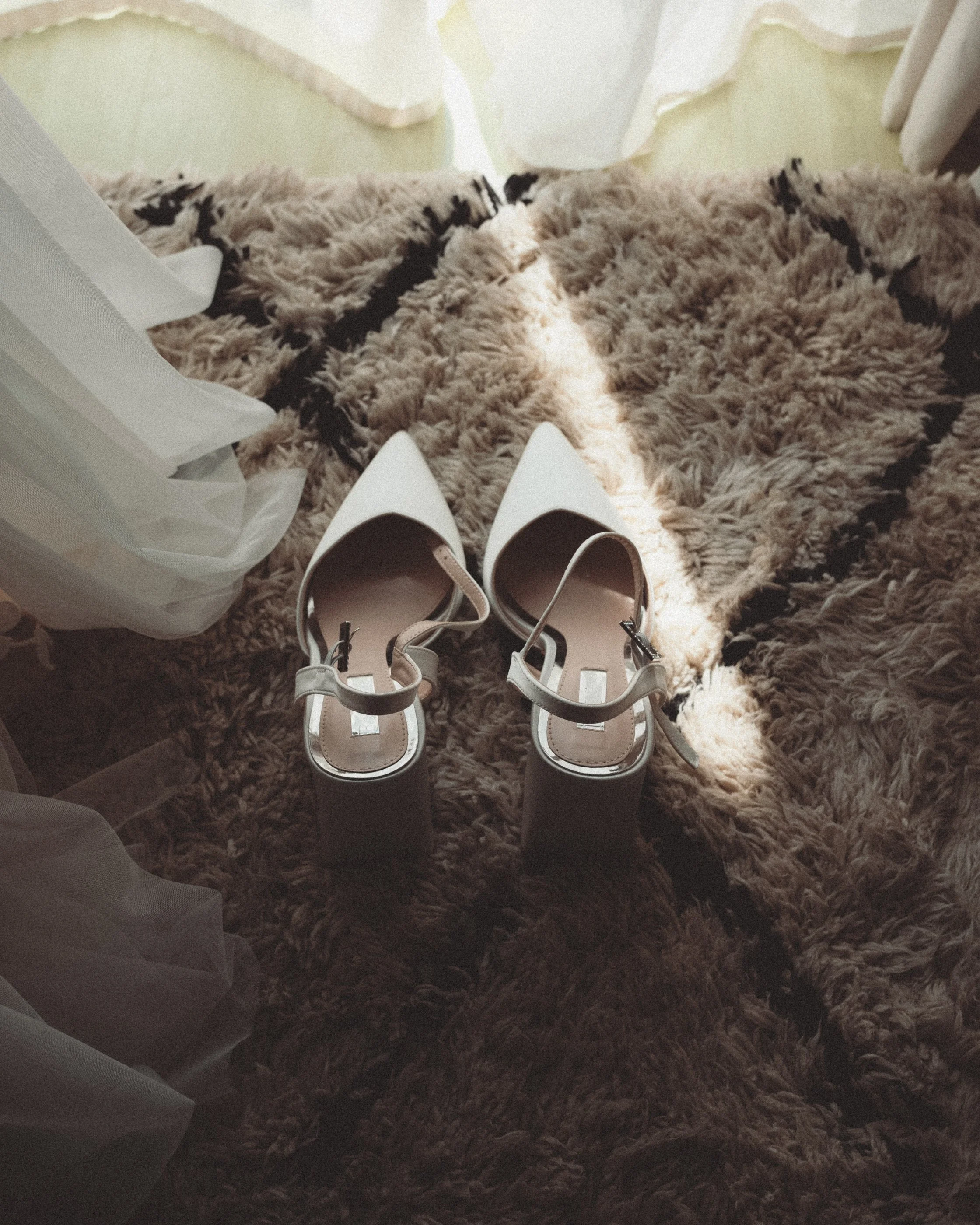Ein Paar weiße High Heels liegt auf einem flauschigen, gemusterten Teppich, daneben eine weiße Einkaufstüte.