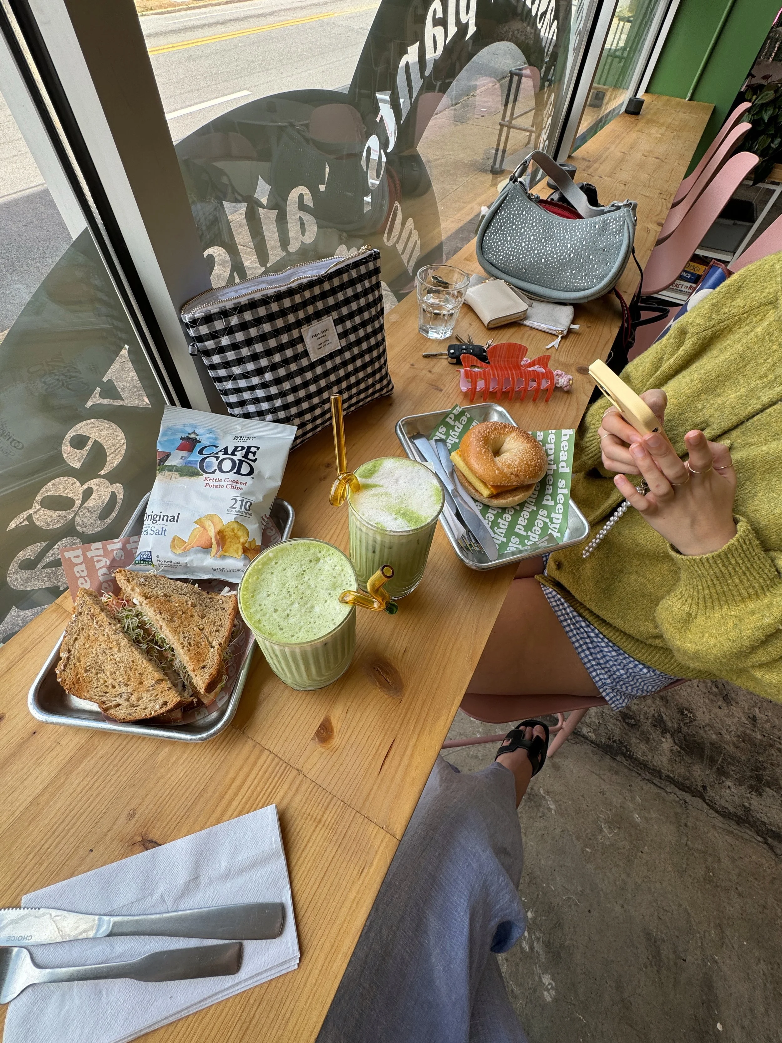 A person sitting at a wooden table in a cafe, holding a smartphone. On the table, there are two green smoothies with straws, a tray with a sandwich, a bag of Cape Cod potato chips, and a tray with a bagel sandwich. Other items on the table include a wallet, a glass of water, a set of keys, a small purse, and a pink hair clip. The cafe has pink chairs, and large windows with an outside view of a street.