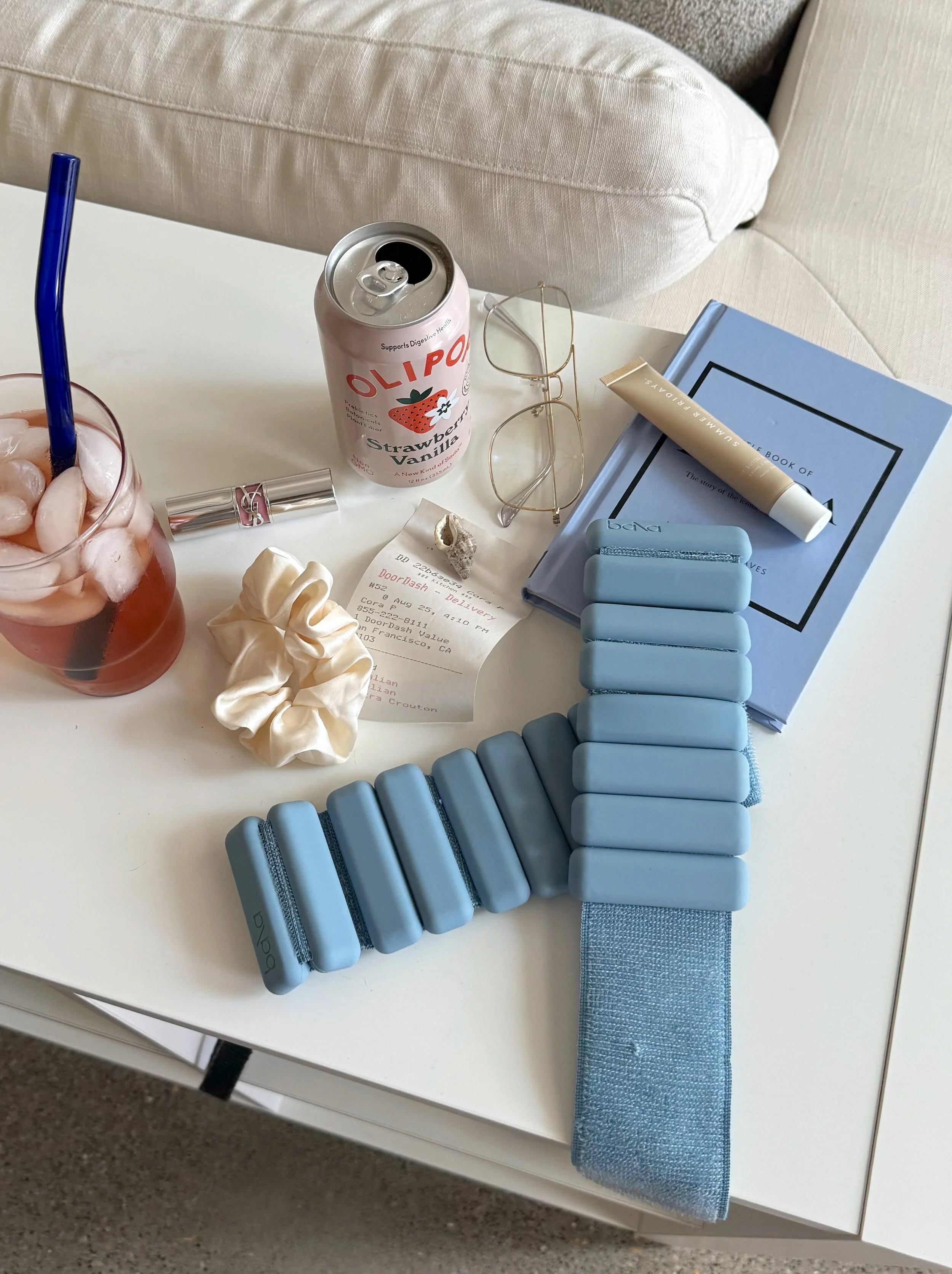 A white table with a blue book, a tube of skin cream, light blue hair clips, a blue ribbon, a crumpled white hair scrunchie, a receipt, a pair of eyeglasses, a can of strawberry vanilla flavored drink, and a pink-colored beverage with ice and a blue straw.
