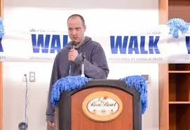 Man speaking at a podium during a walk event, with a banner that says 'WALK' behind him, and decorations on the podium.