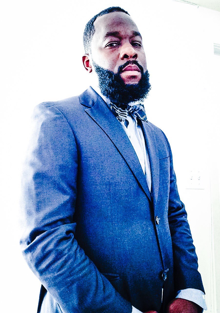 A man with a beard wearing a dark blue suit, light dress shirt, and patterned tie standing against a plain white background.