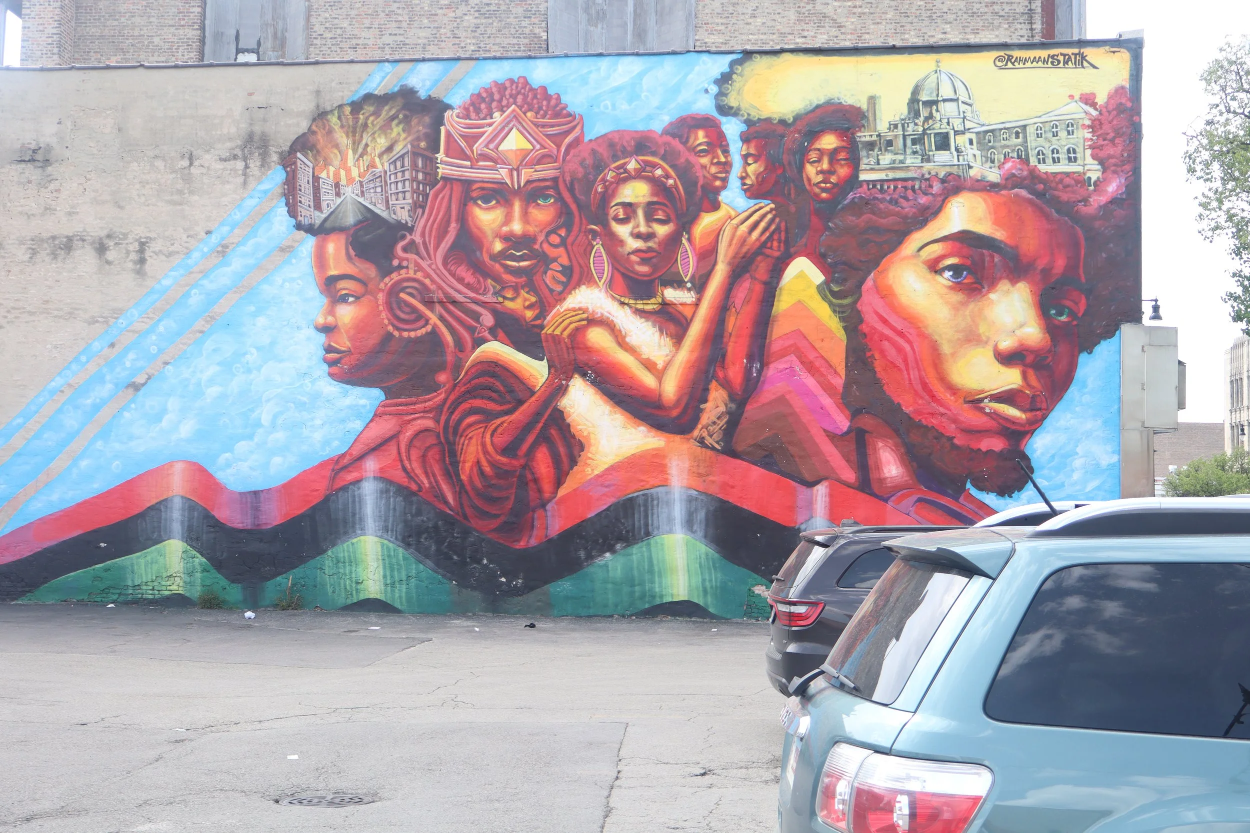 Colorful mural depicting diverse faces of Black women, some with traditional headpieces, with buildings and natural elements integrated into the artwork, located on a city wall.