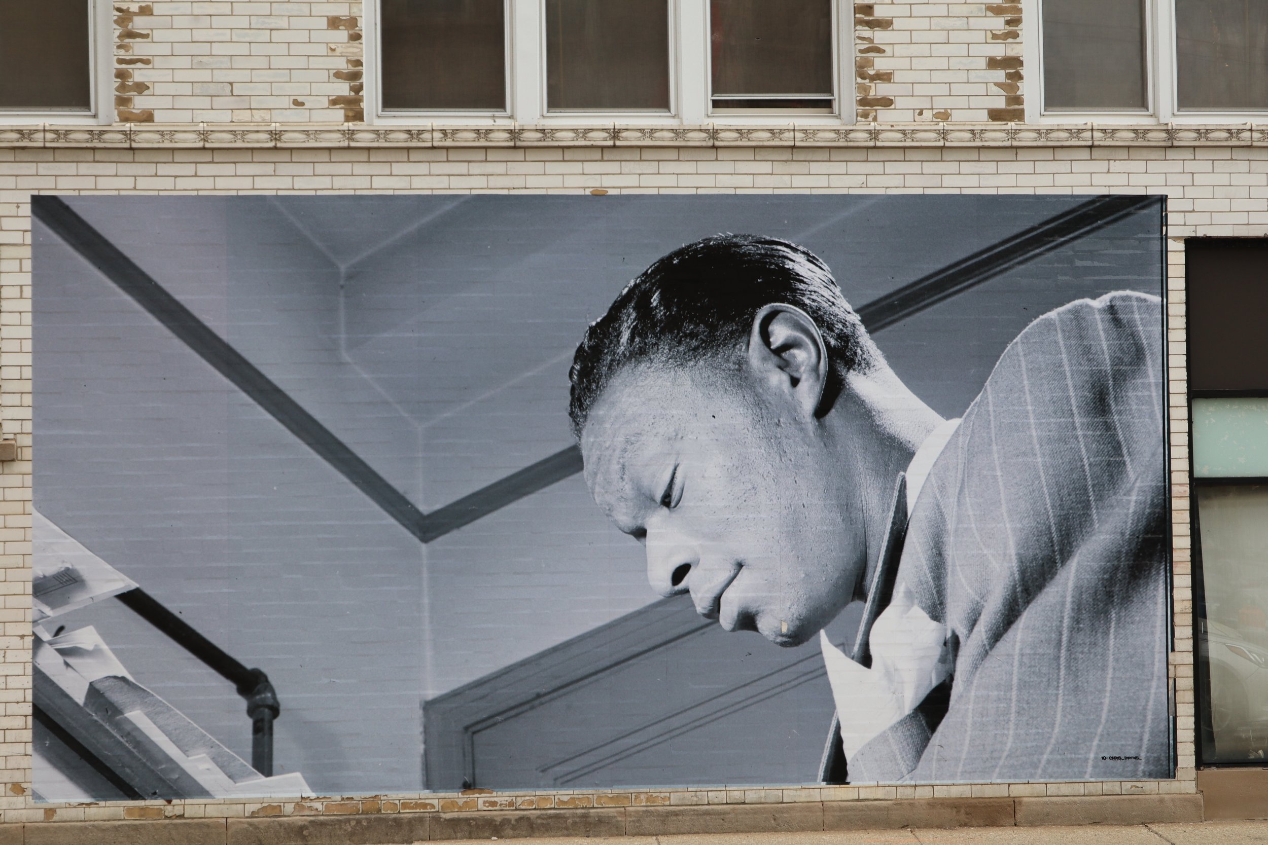 Large black and white mural of a woman playing the piano, focusing on her face and hands, on the side of a brick building with windows above.