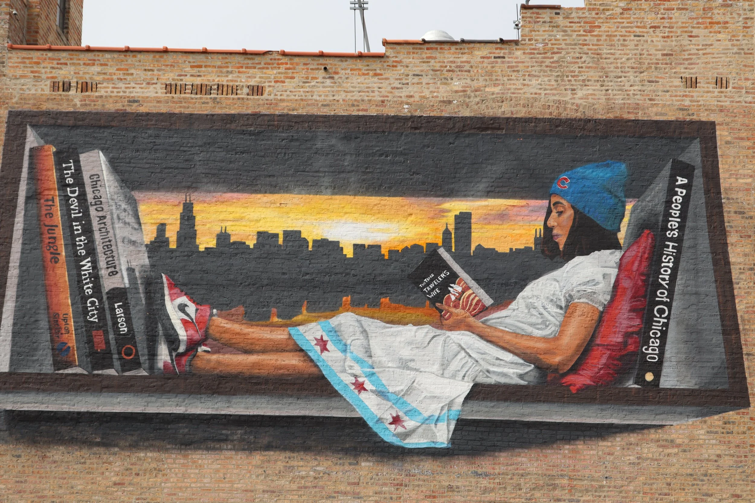 A mural on a brick wall depicting a woman with dark hair wearing a blue Chicago Cubs baseball cap, reading a newspaper with the headline about the history of Chicago.