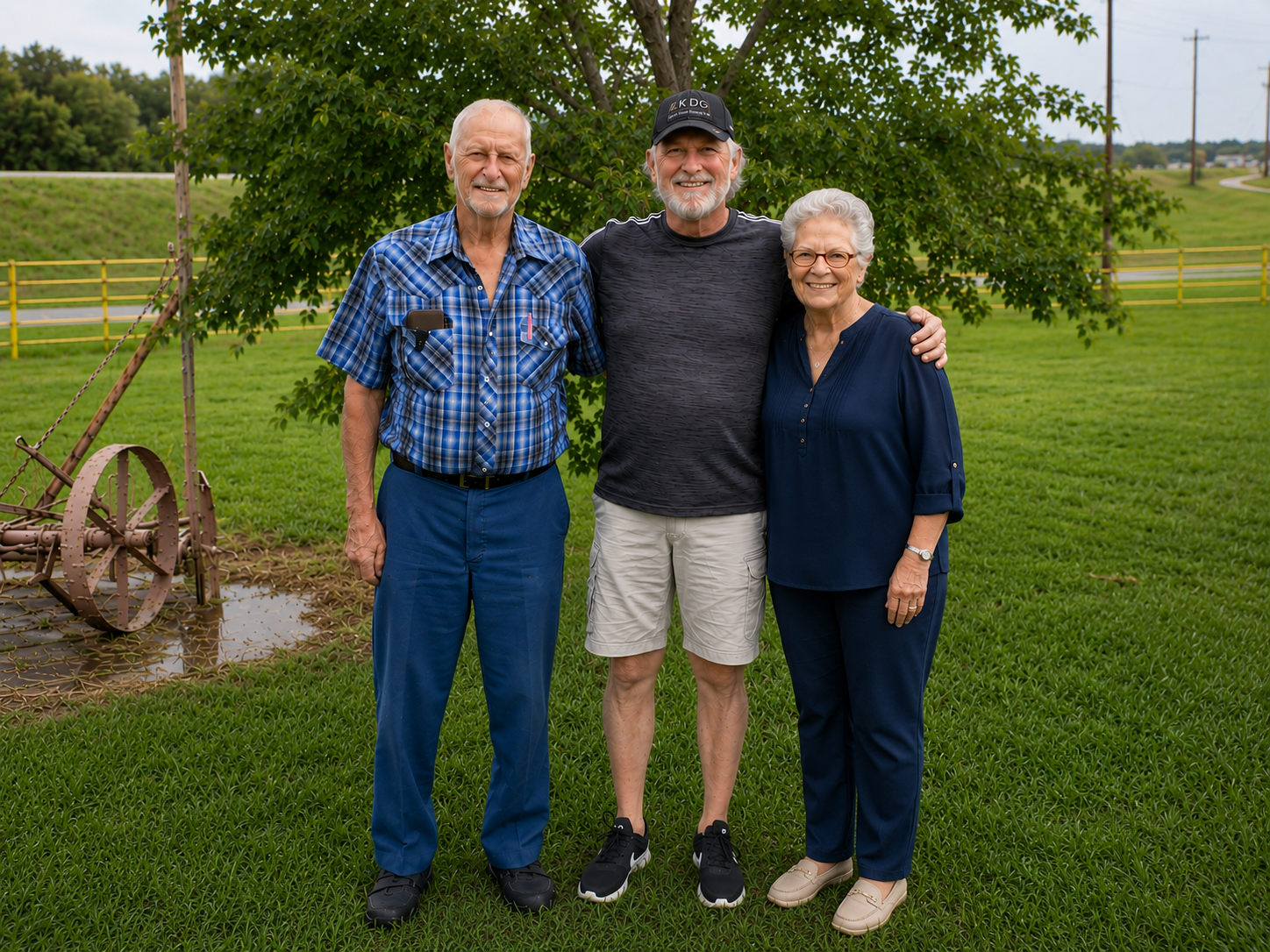 Three elderly adults, two men and one woman, standing outdoors on a grassy area with a tree and a wooden fence in the background, smiling and posing for a photo.