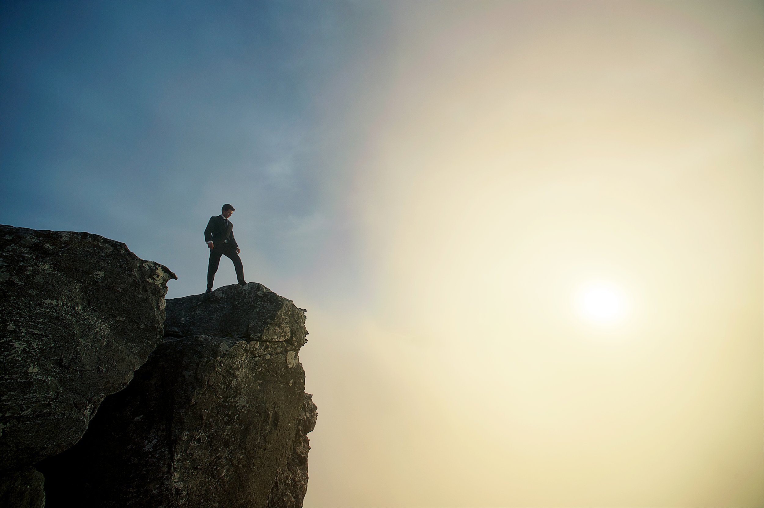 A man in a suit walking along a rocky cliff edge with the sky and sunset in the background.