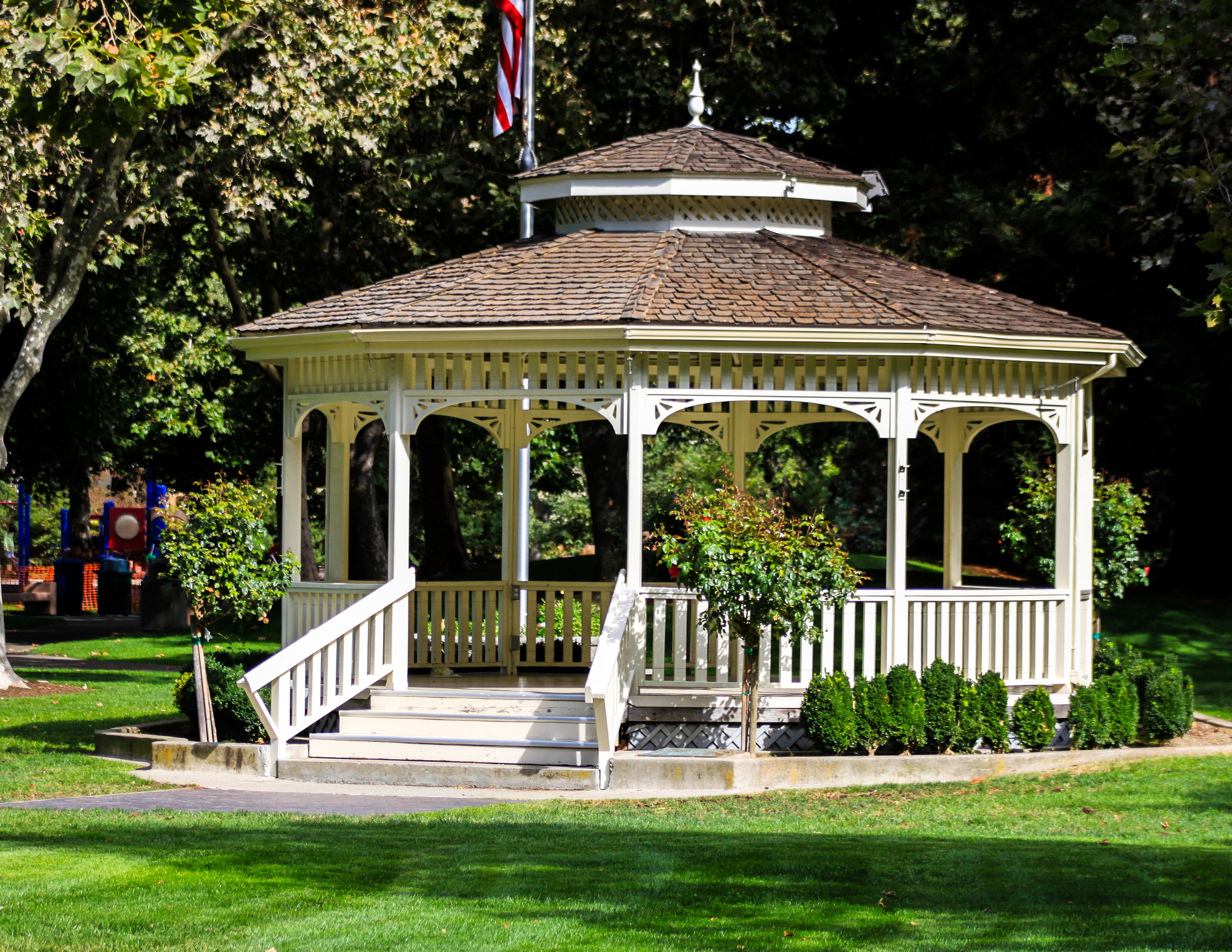 A white wooden gazebo with a shingled roof and ornate trim, situated in a park with green grass and trees in the background, an American flag on a pole nearby.