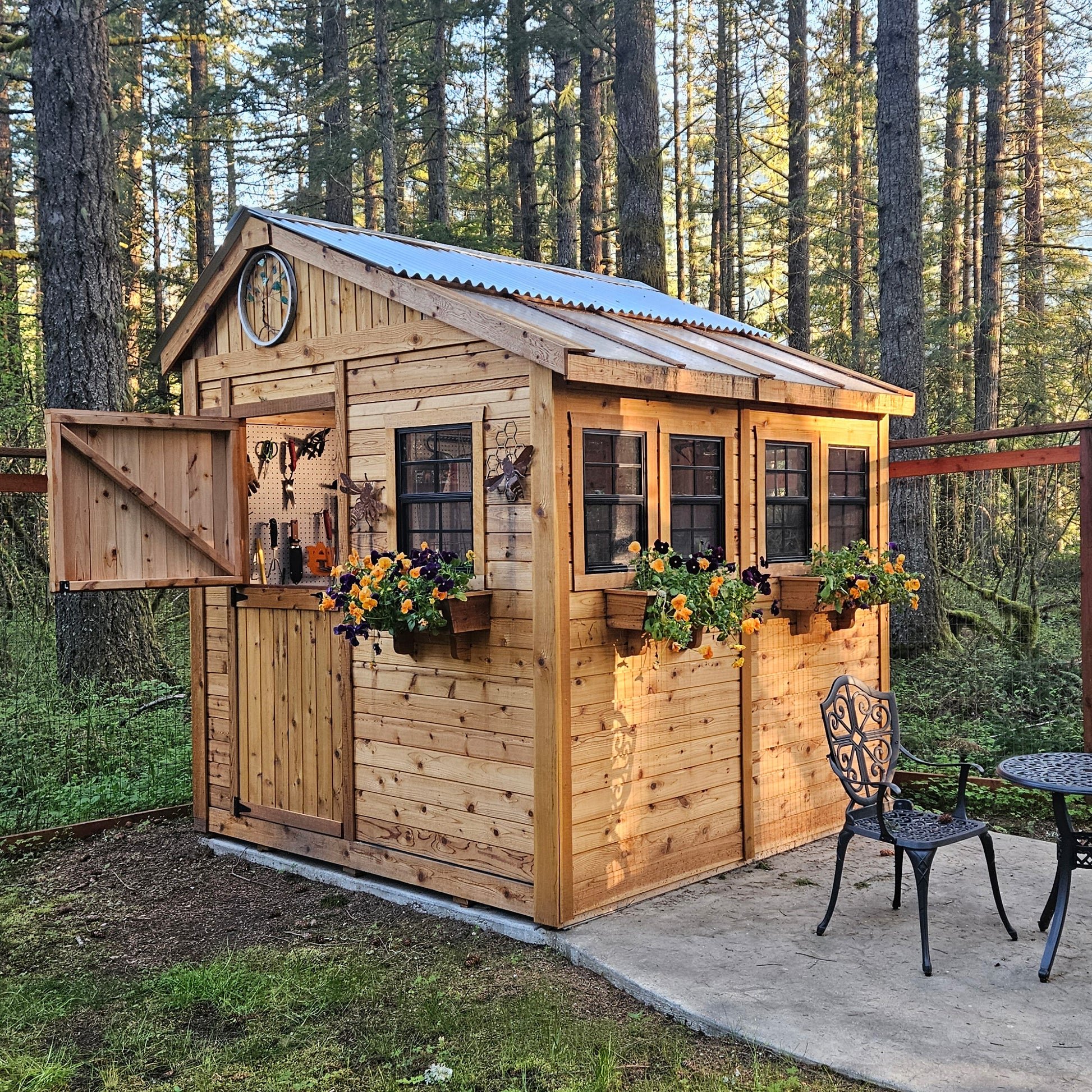 A small wooden shed with a metal roof, surrounded by trees, with flower boxes attached to the outside and a decorative clock on the upper corner.