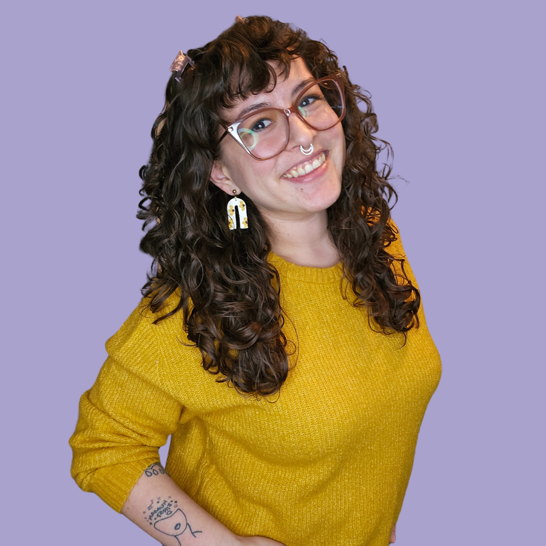 A young woman with curly brown hair, glasses, and multiple piercings, smiling while wearing a yellow sweater against a purple background.