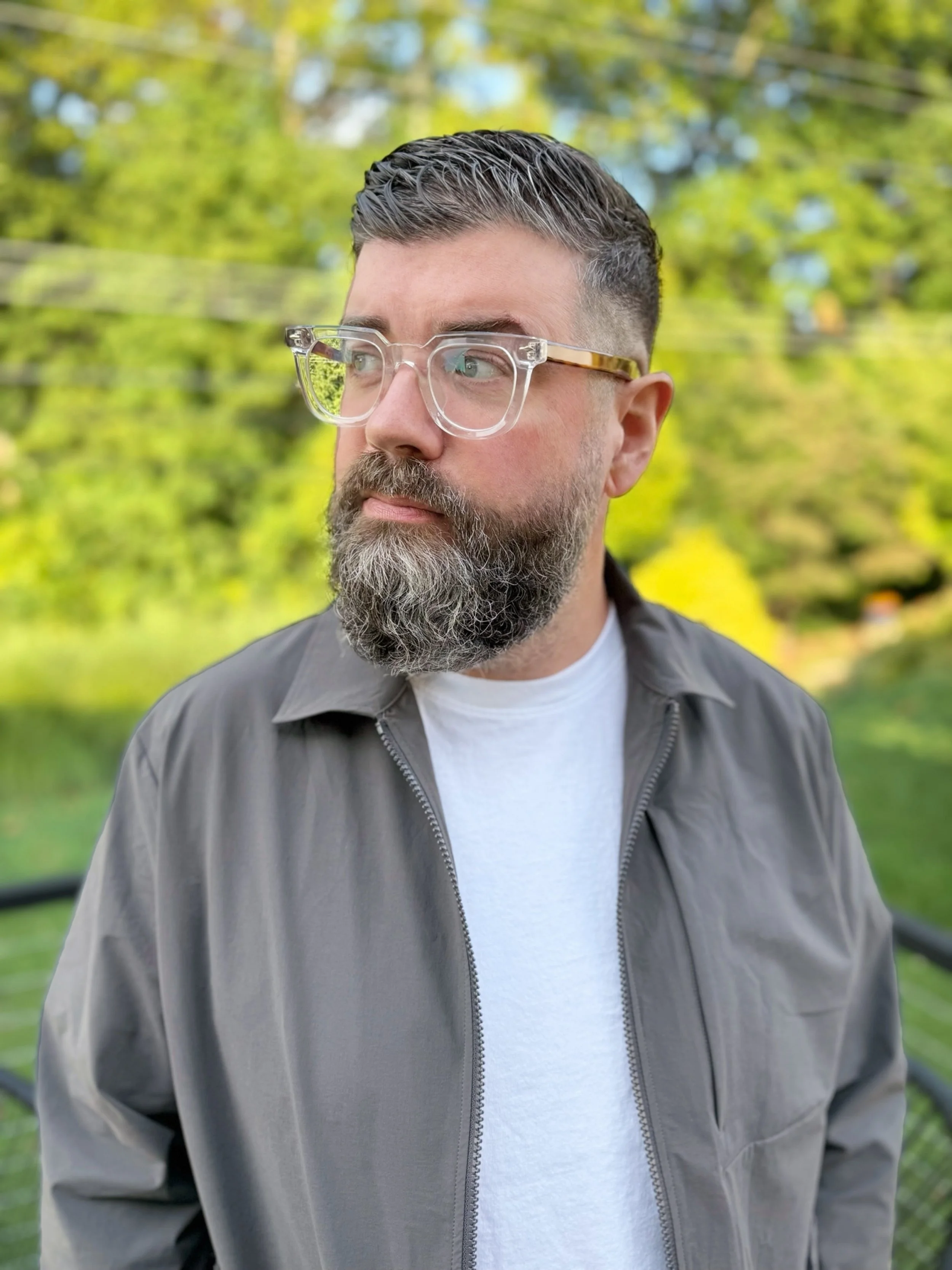 A man with glasses and a well-groomed beard is outdoors, with green trees in the background. He is wearing a gray jacket over a white shirt and looking to the left.