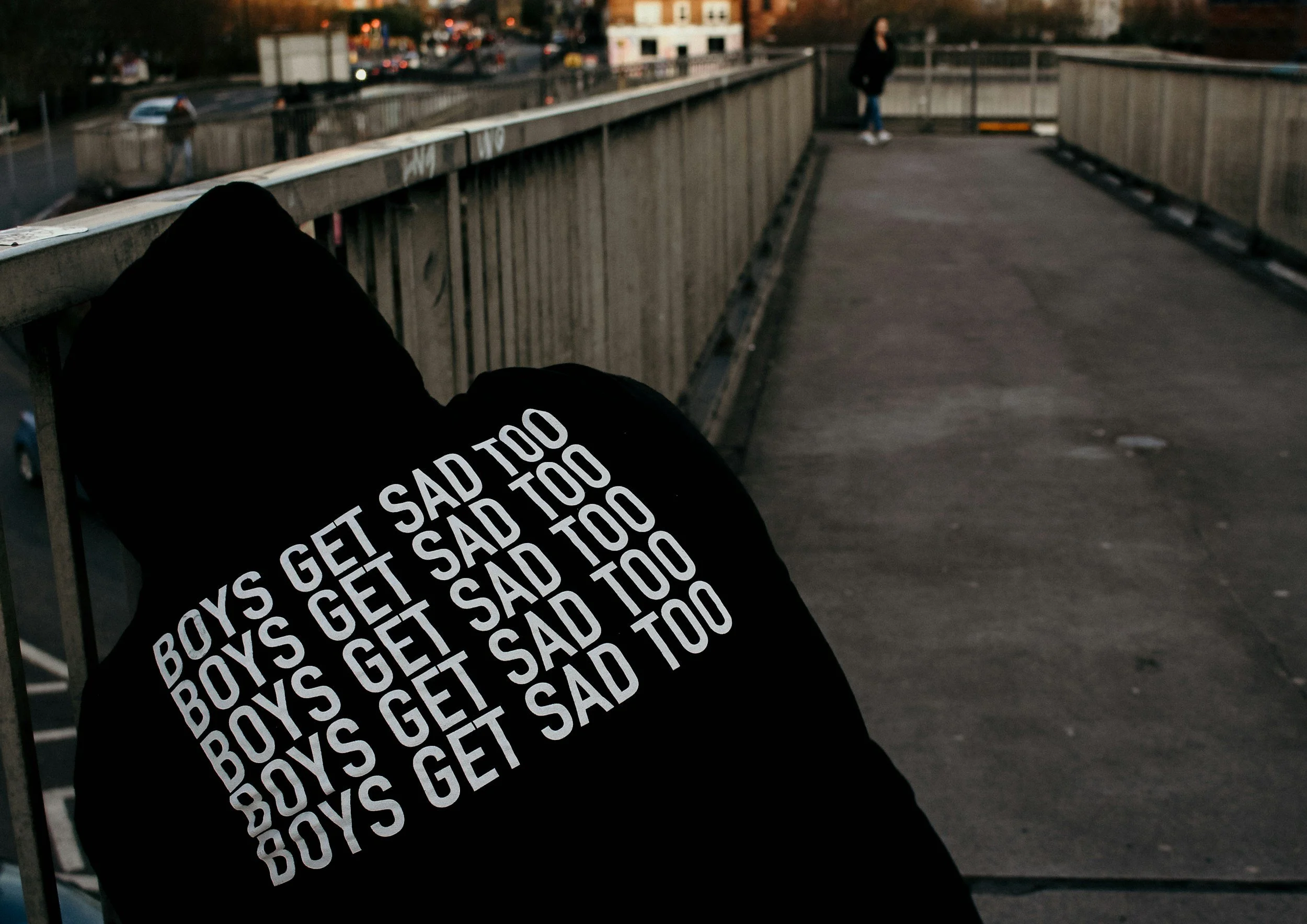Person sitting on a bench on a bridge, wearing a black hoodie with repeated white text reading 'GET SAD TOO'. In the background, another person is standing at the end of the bridge, and city buildings and traffic are visible.