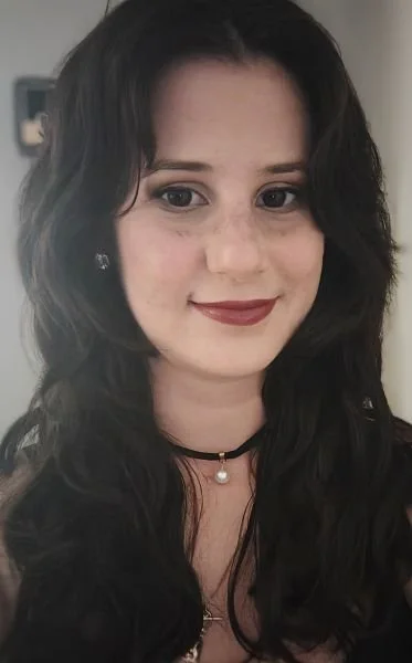 A woman with long, dark, wavy hair and light makeup, smiling at the camera, wearing earrings and a choker with a pearl pendant.