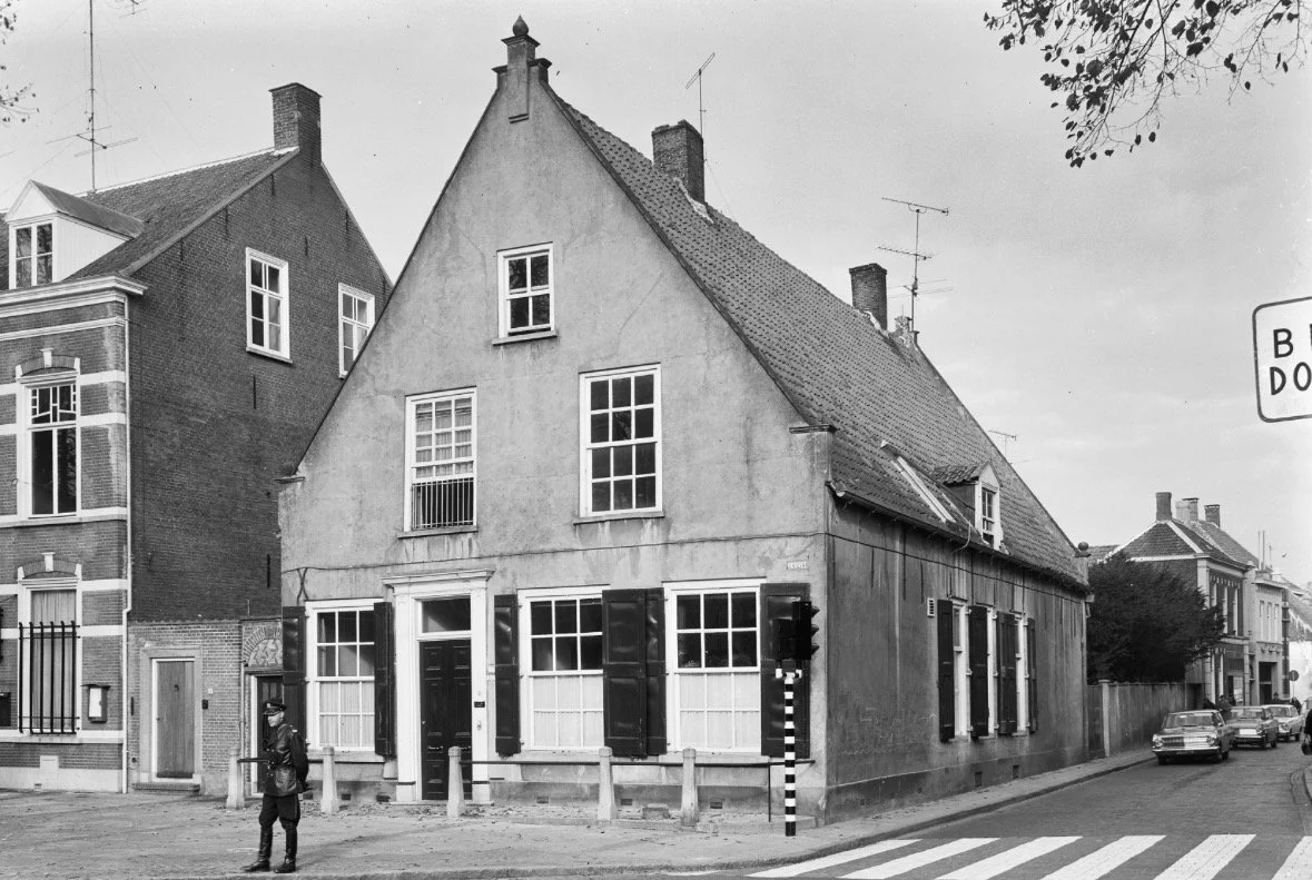 Een zwart-wit foto van een oud huis met een steil dak, meerdere ramen met luiken, en een deur met een wit raam. Een man staat op de straat, en er zijn enkele auto's en andere gebouwen op de achtergrond.