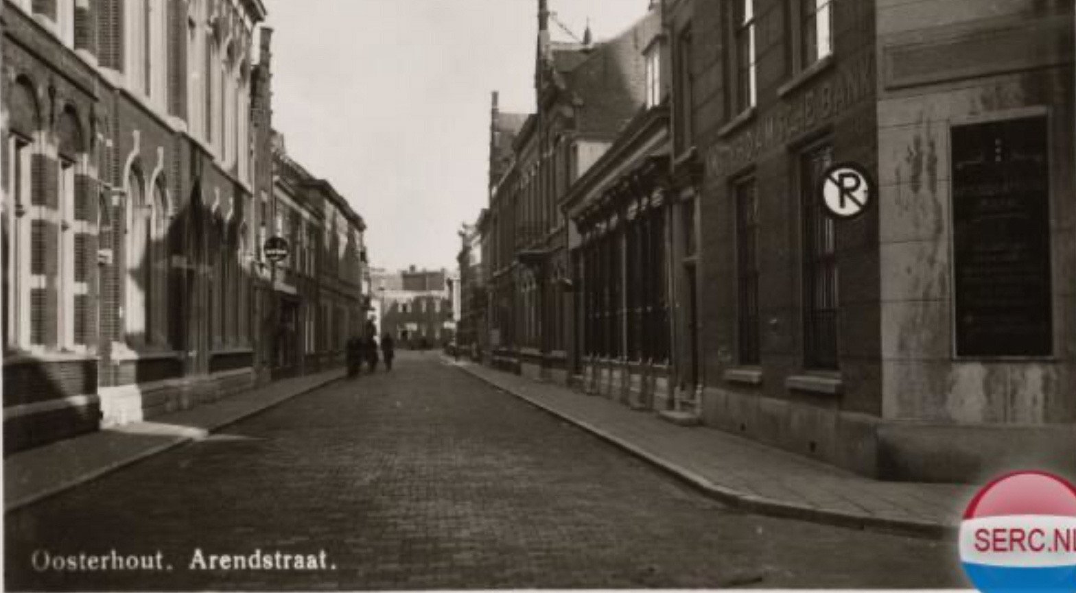 Een oude zwart-wit foto van een straat in Oosterhout met historische gebouwen aan beide zijden, een kasseienweg en enkele mensen wandelend in de verte.