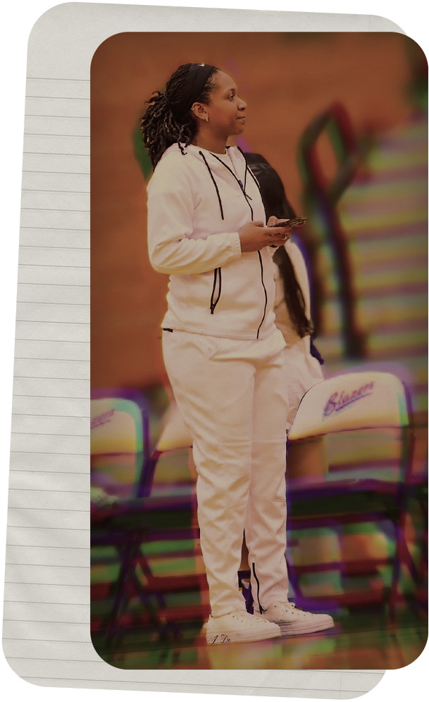 A woman in a white hoodie and sweatpants stands in a gym, holding a phone, with chairs and a colorful wall in the background.