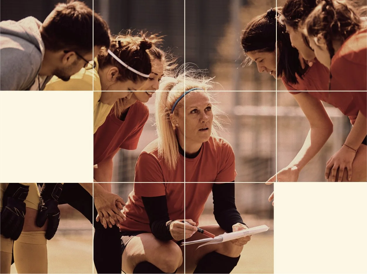 A female coach with blonde hair and wearing a red sports shirt is kneeling on a sports field, holding a clipboard, and speaking to a group of young female athletes who are bent over listening attentively.