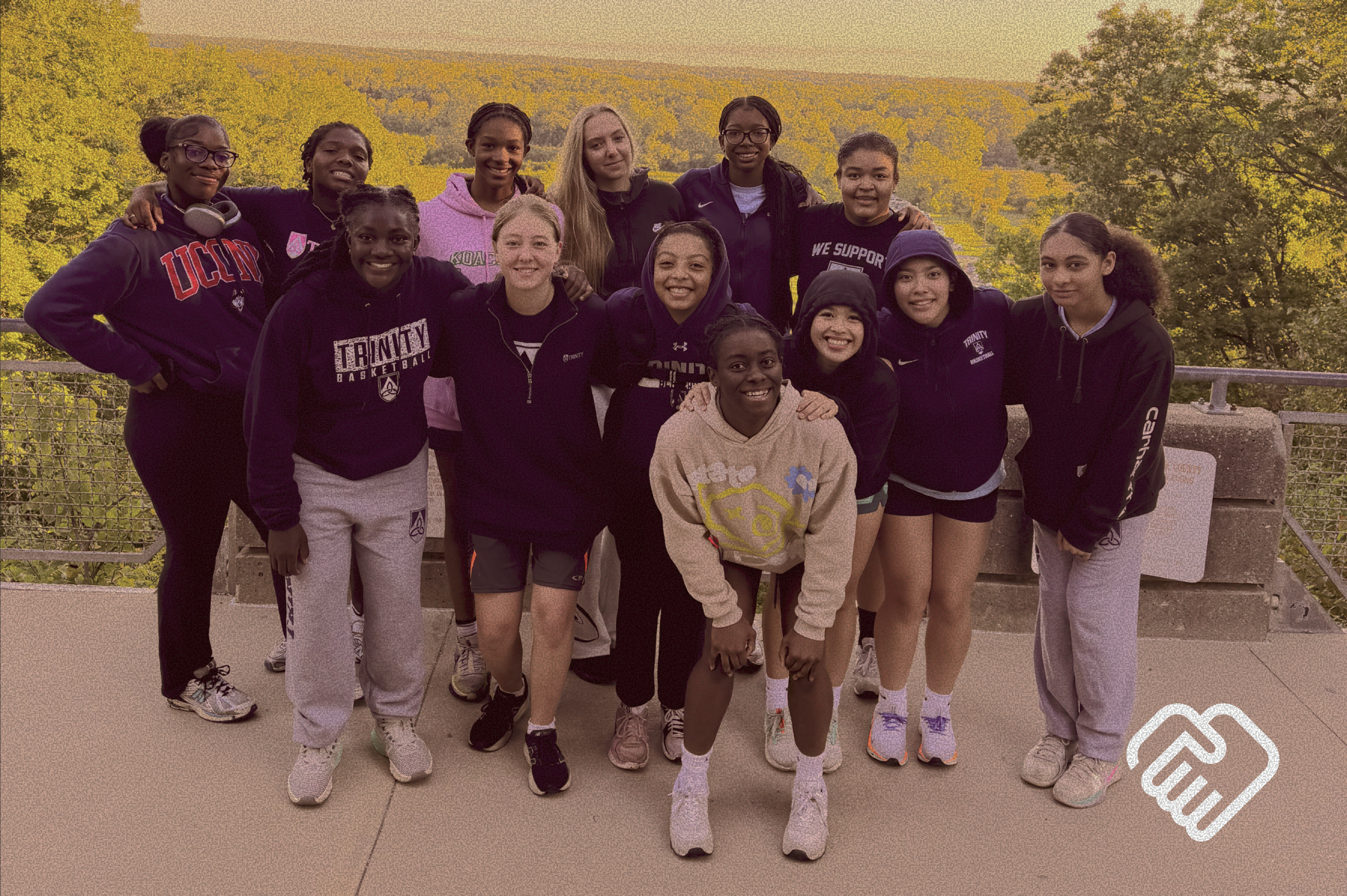 A group of young women and girls standing together outdoors on a railing with trees and a sunset in the background. They are smiling and wearing casual sportswear.
