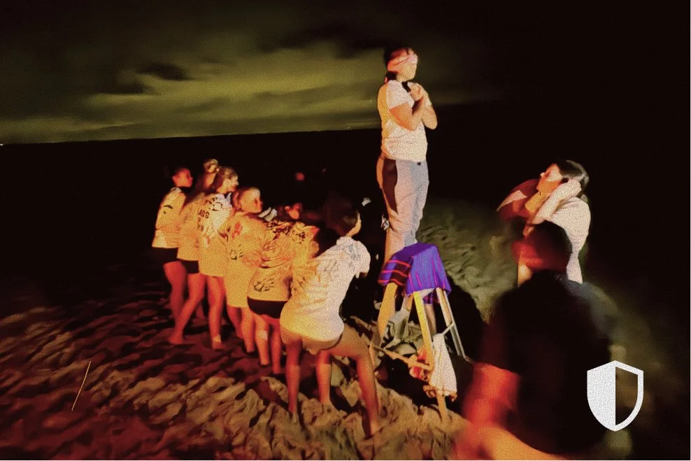 A group of children and adults are gathered around a person standing on a chair on a beach at night, performing a trust fall exercise.