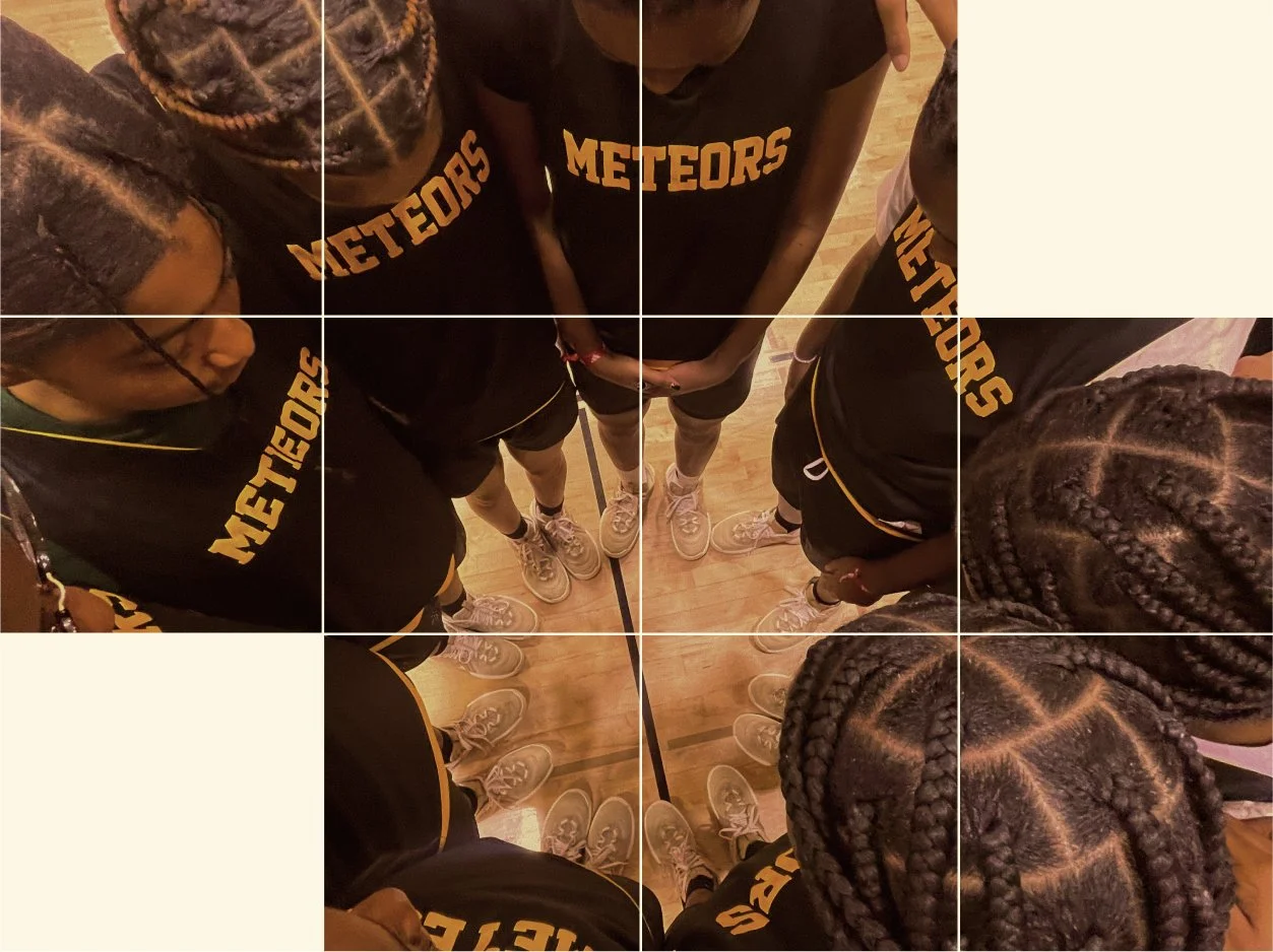 Birds-eye view of a group of young women in black basketball uniforms with yellow lettering that says 'METEORS,' standing in a circle on a basketball court, looking down at the ground together.
