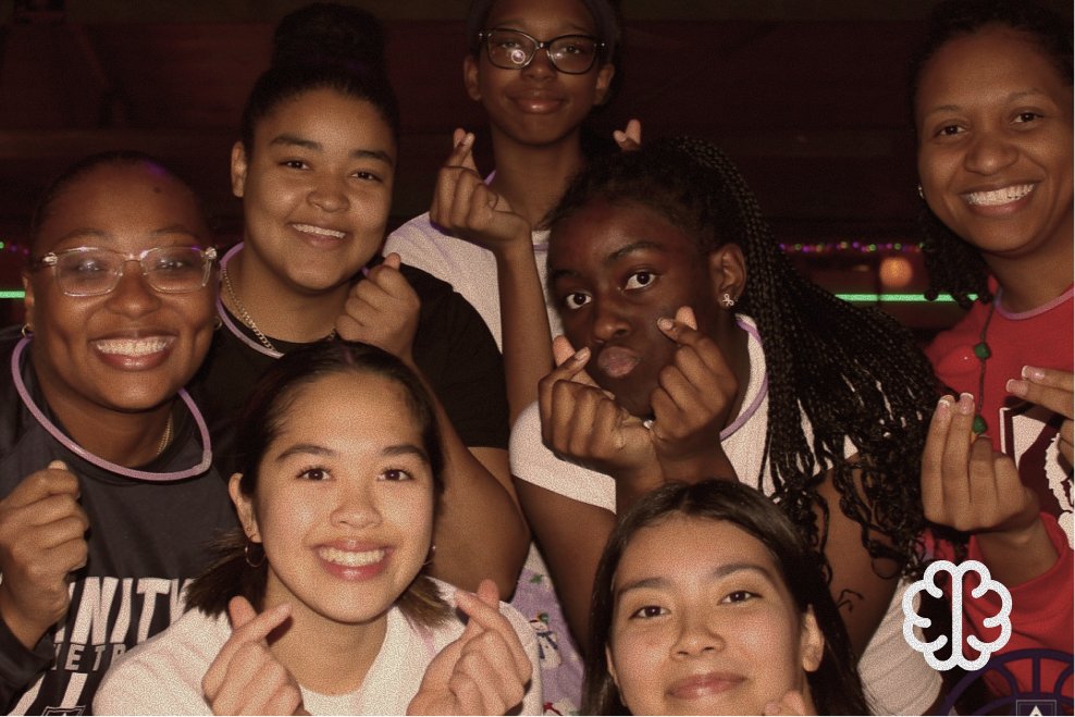 Group of seven young women smiling and making hand gestures, posing for a photo in a dimly lit room.