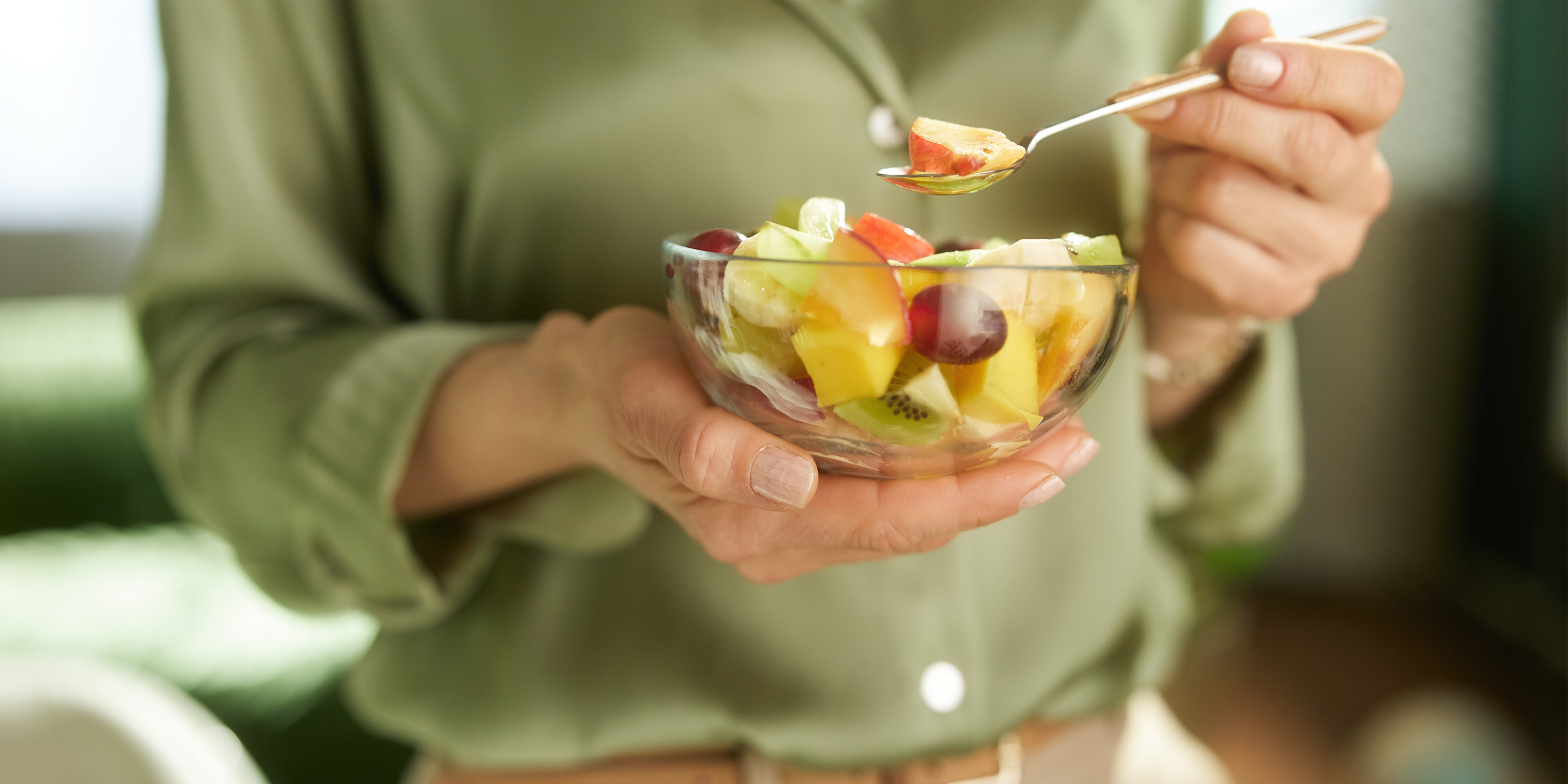 A person in a green sweater holds a glass bowl filled with assorted chopped fruits and uses a fork to pick up a piece of fruit.