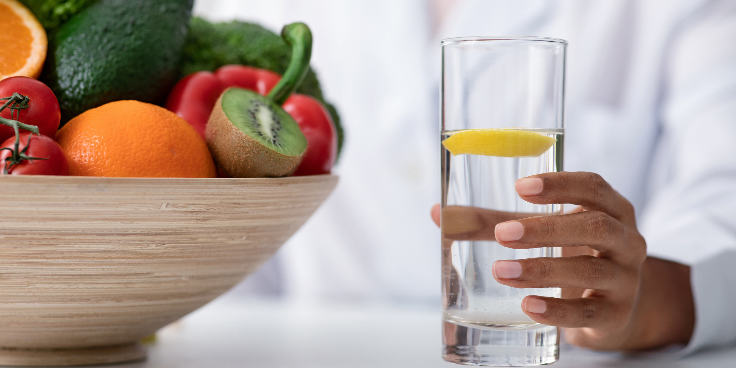 A person holding a glass of water with a lemon slice, next to a wooden bowl filled with various fruits including an orange, tomatoes, an avocado, an apple, a kiwi, and an orange half.