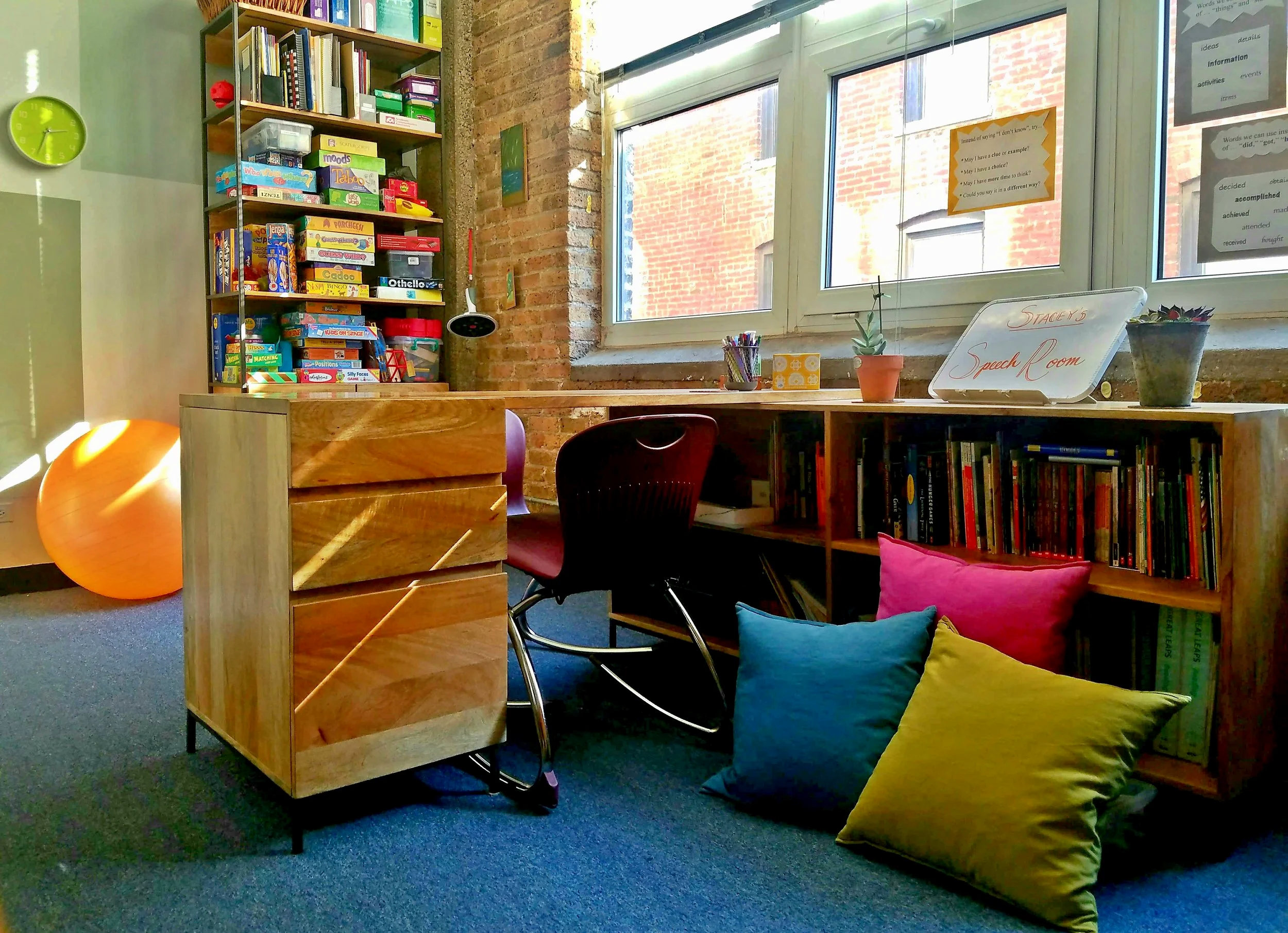 A cozy reading nook with a wooden desk, colorful pillows on the floor, a bookshelf filled with games and books, a window letting in natural light, and plants and decorative items on the desk.