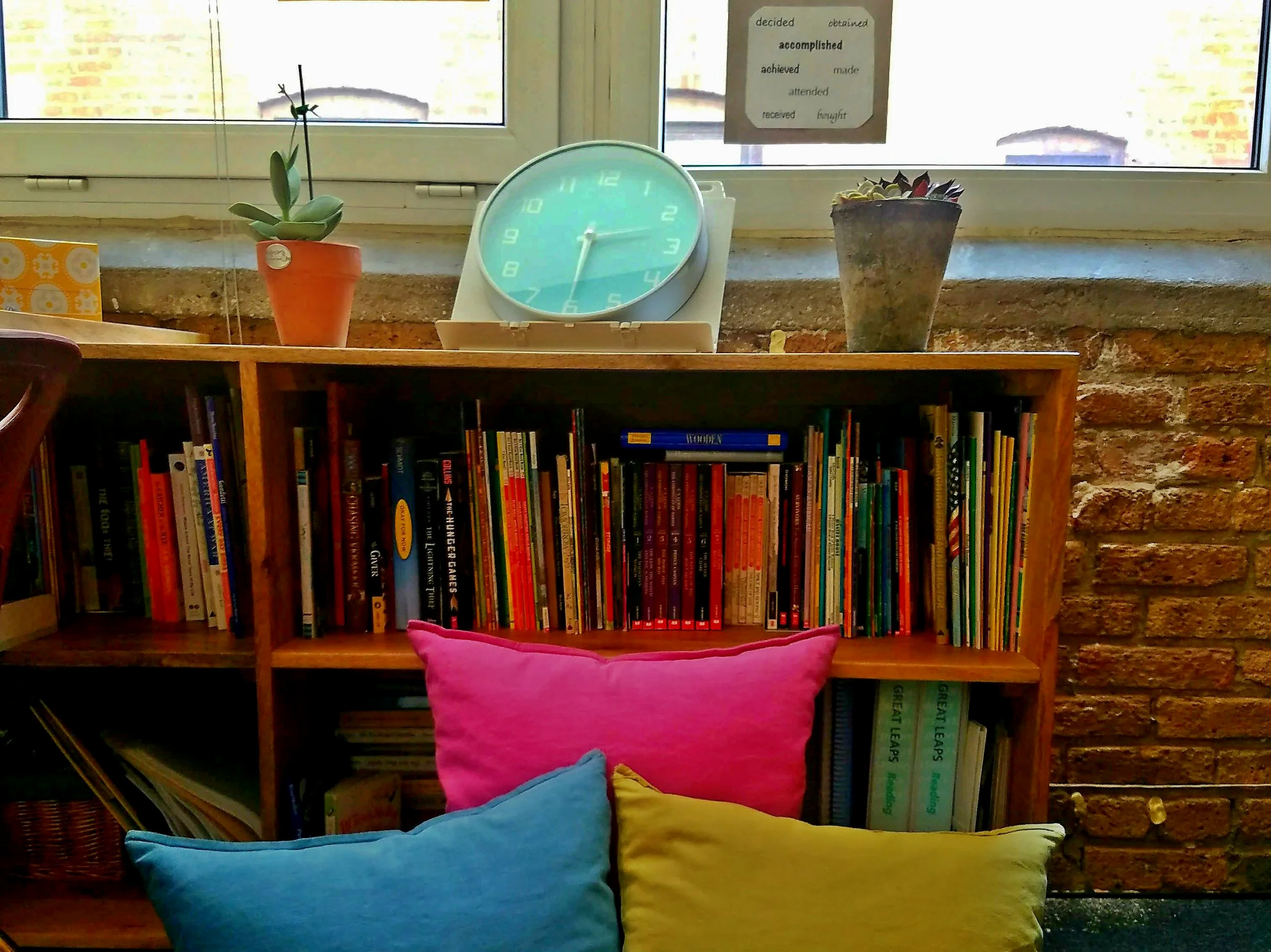 A bookshelf filled with colorful books underneath a window. On top of the bookshelf are two potted plants, one with green leaves and another with dark purple leaves. A large pink, yellow, and blue cushion rests on the floor in front of the bookshelf. A clock showing 1:15 is placed on the window sill. There is a paper with words like 'decided,' 'accomplished,' 'achieved,' 'made,' 'attended,' 'received,' and 'brought' taped to the window.
