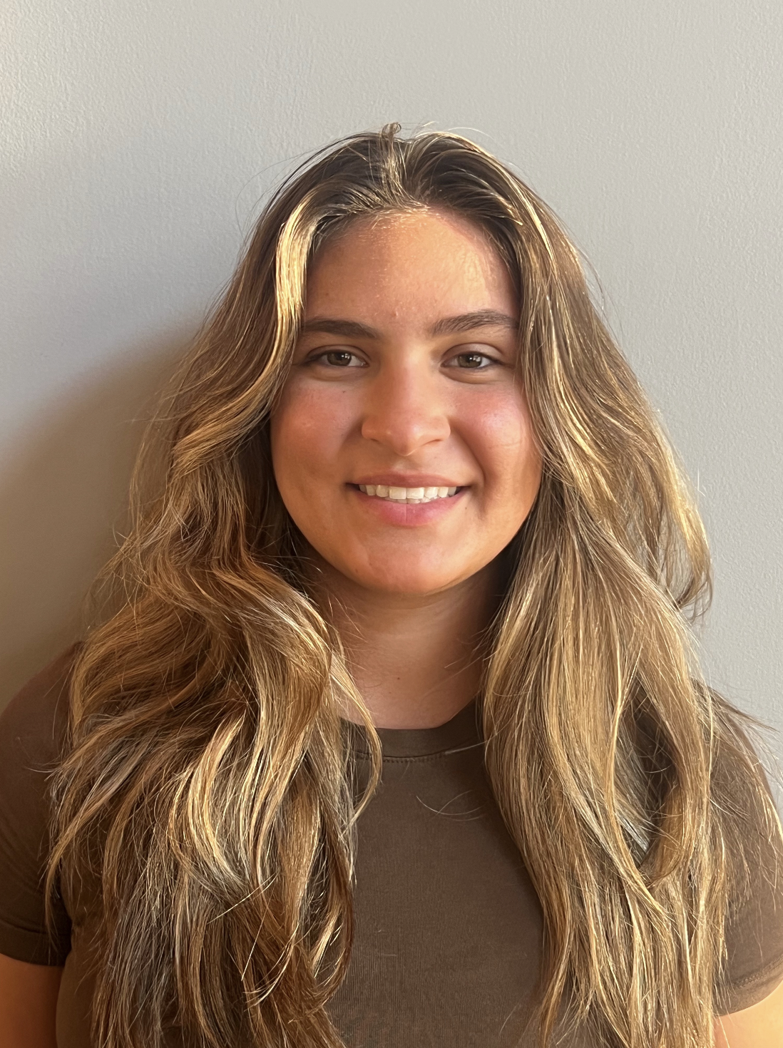 A young woman with long, wavy blonde hair smiling in front of a plain beige wall.