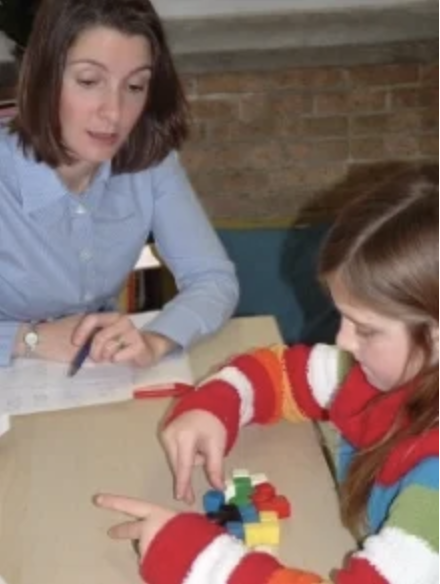 A woman and a girl are sitting at a table, with the girl playing with colorful building blocks. The woman looks on, possibly giving guidance or instructions.