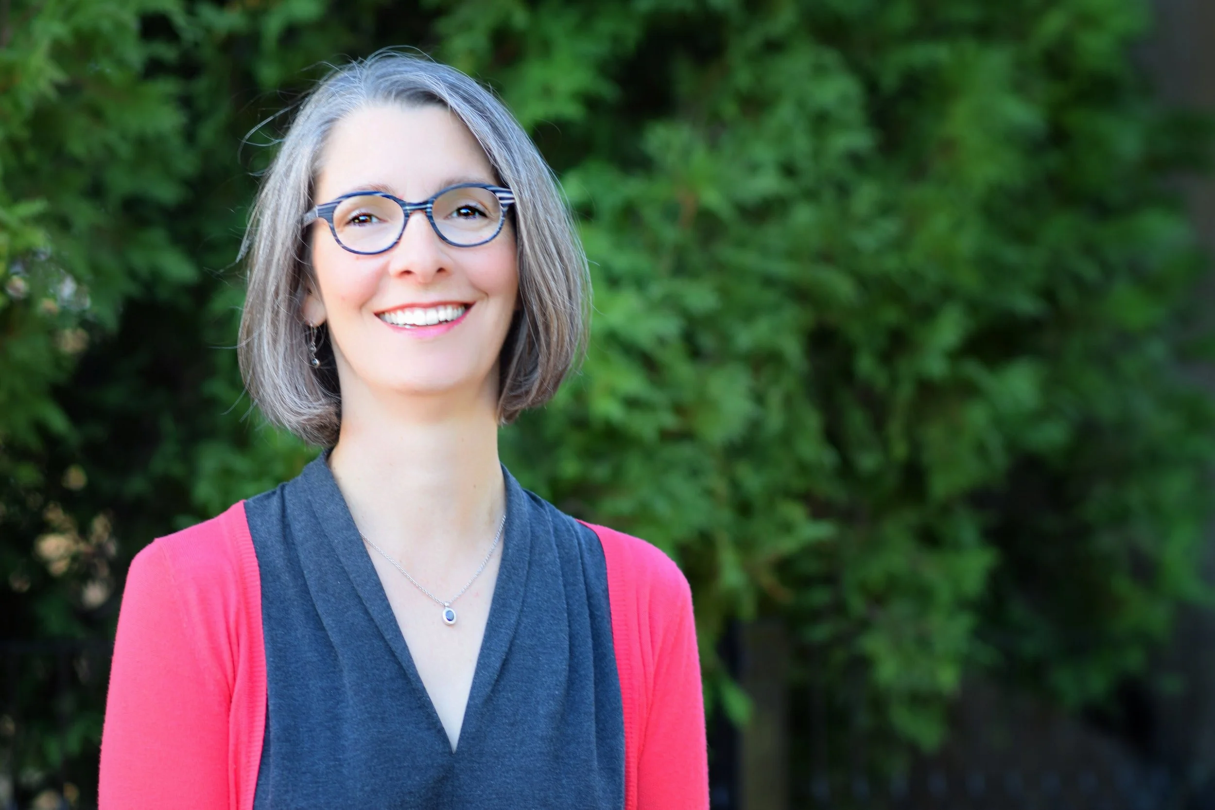 A smiling woman with gray hair and glasses standing outdoors near green foliage, wearing a pink and gray outfit.