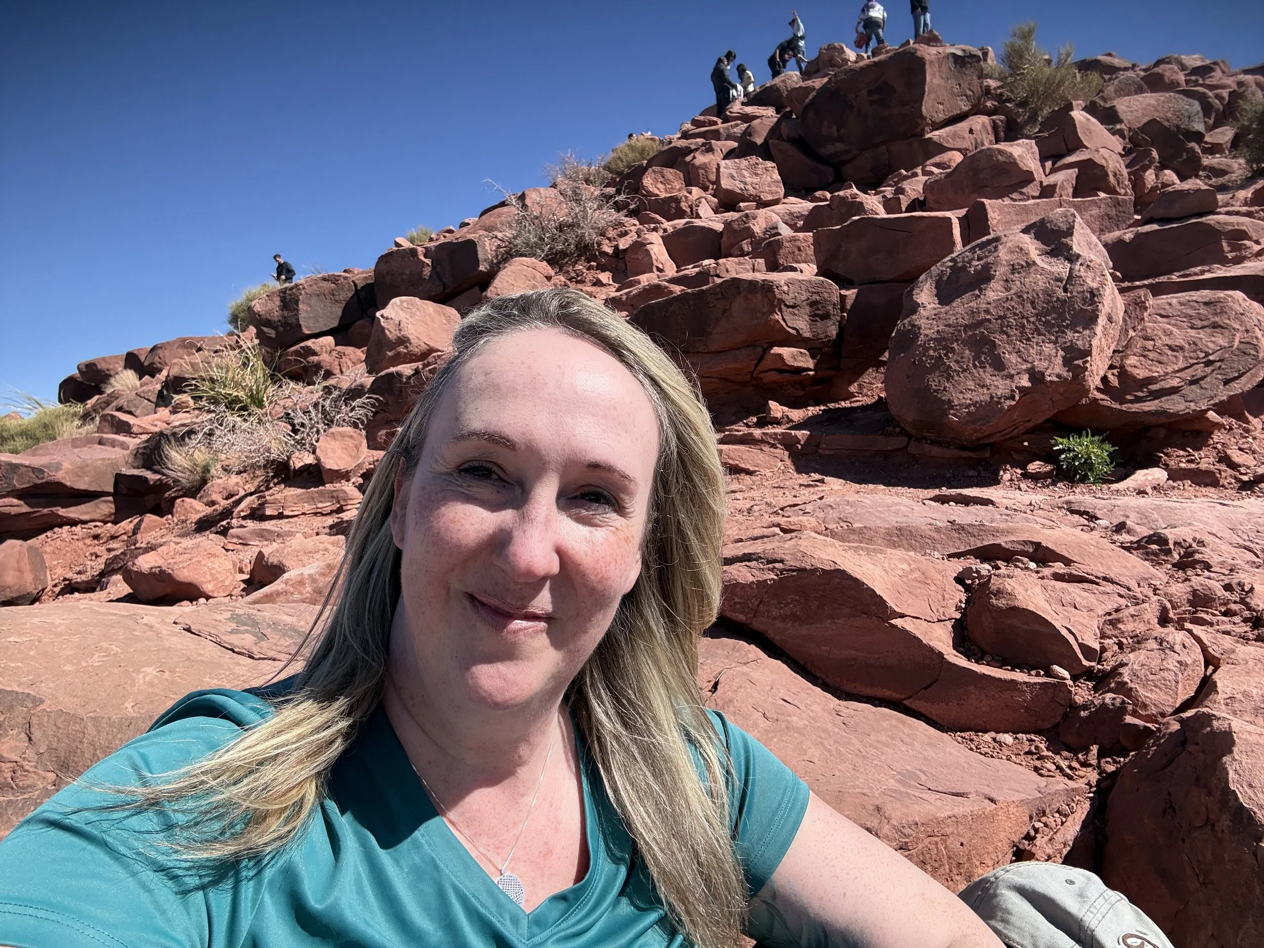 A woman taking a selfie outdoors on a rocky terrain with reddish rocks and sparse desert vegetation, with a clear blue sky overhead. There are several people climbing or standing on the rocks in the background.