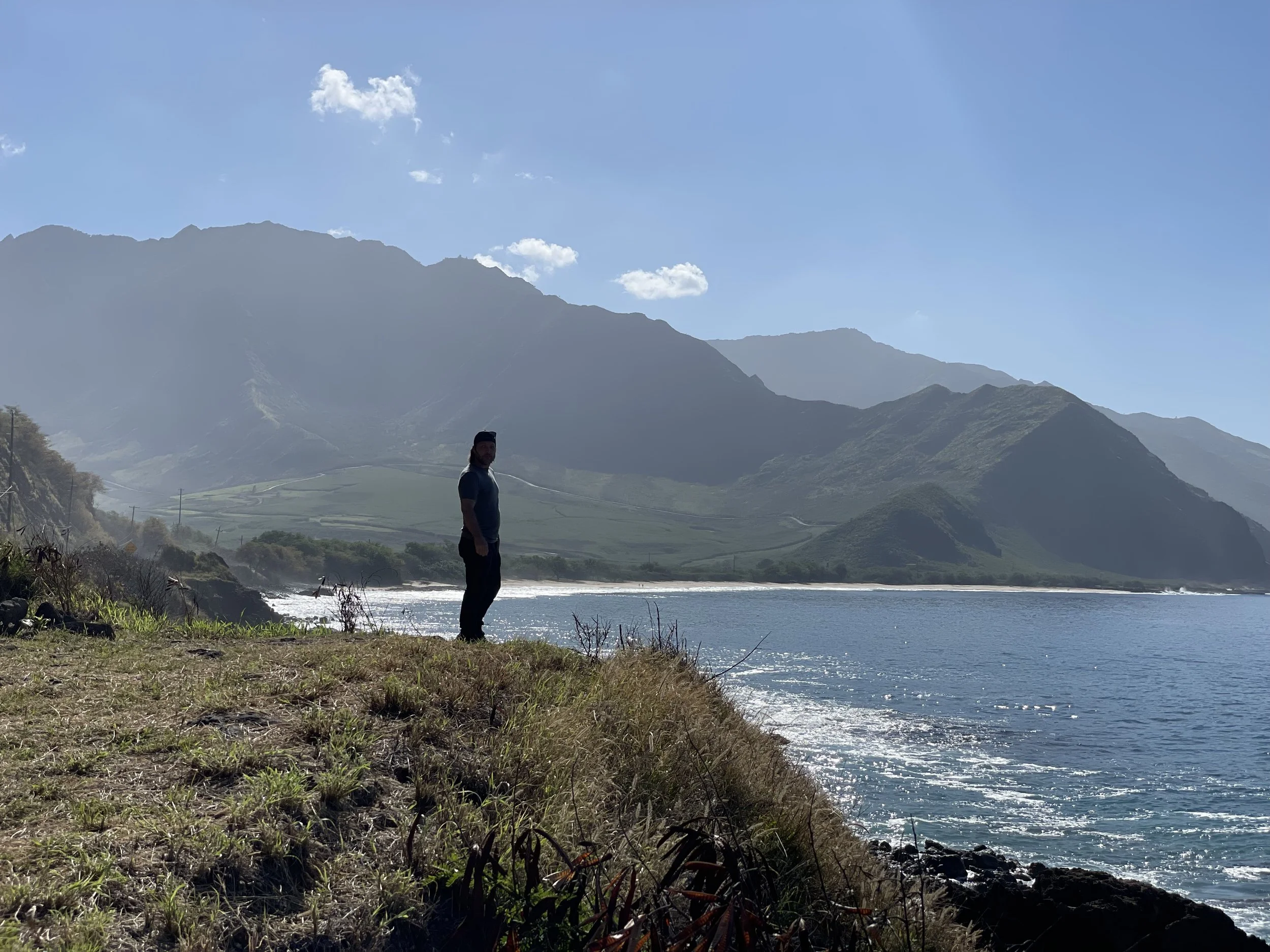 A man standing on the grassy shoreline of Hawaii beaches, with mountains in the background under a clear blue sky.