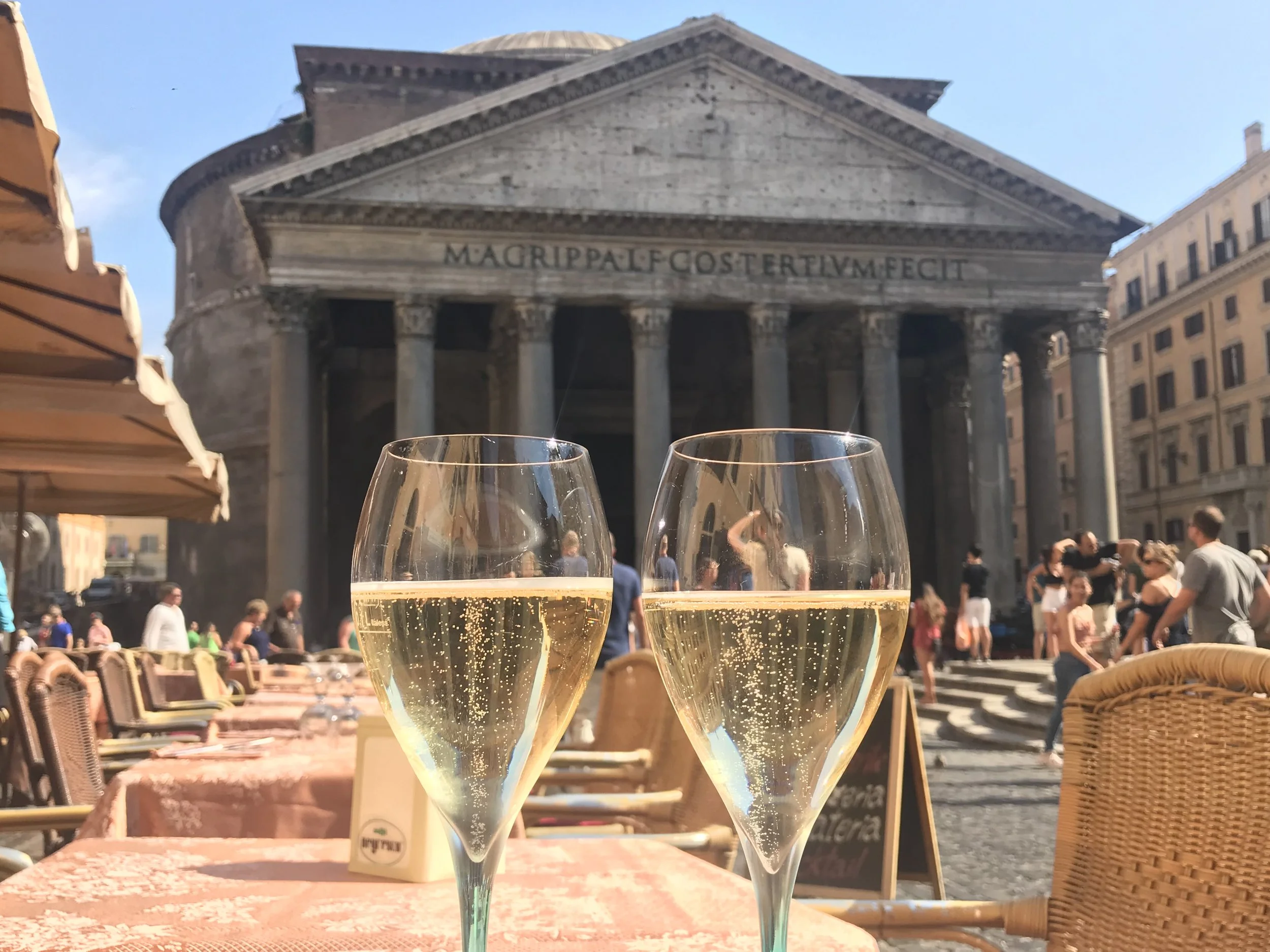 Two glasses of sparkling wine on a table at an outdoor cafe with people gathered in front of an ancient Roman temple, the Pantheon, in Rome, Italy.