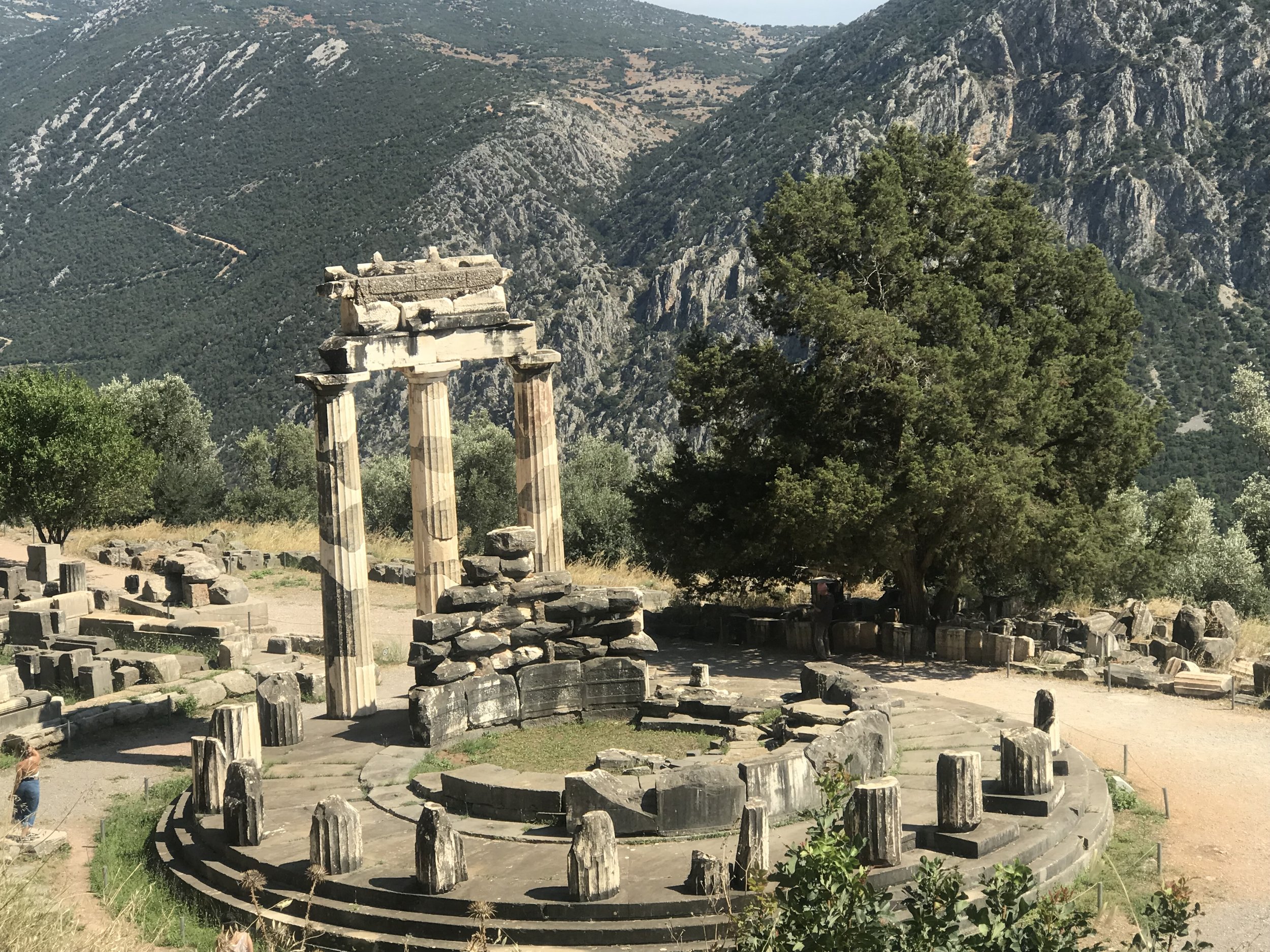 Ancient Greek ruins with three standing columns and scattered stone fragments, situated in a mountainous landscape with trees and a clear sky.
