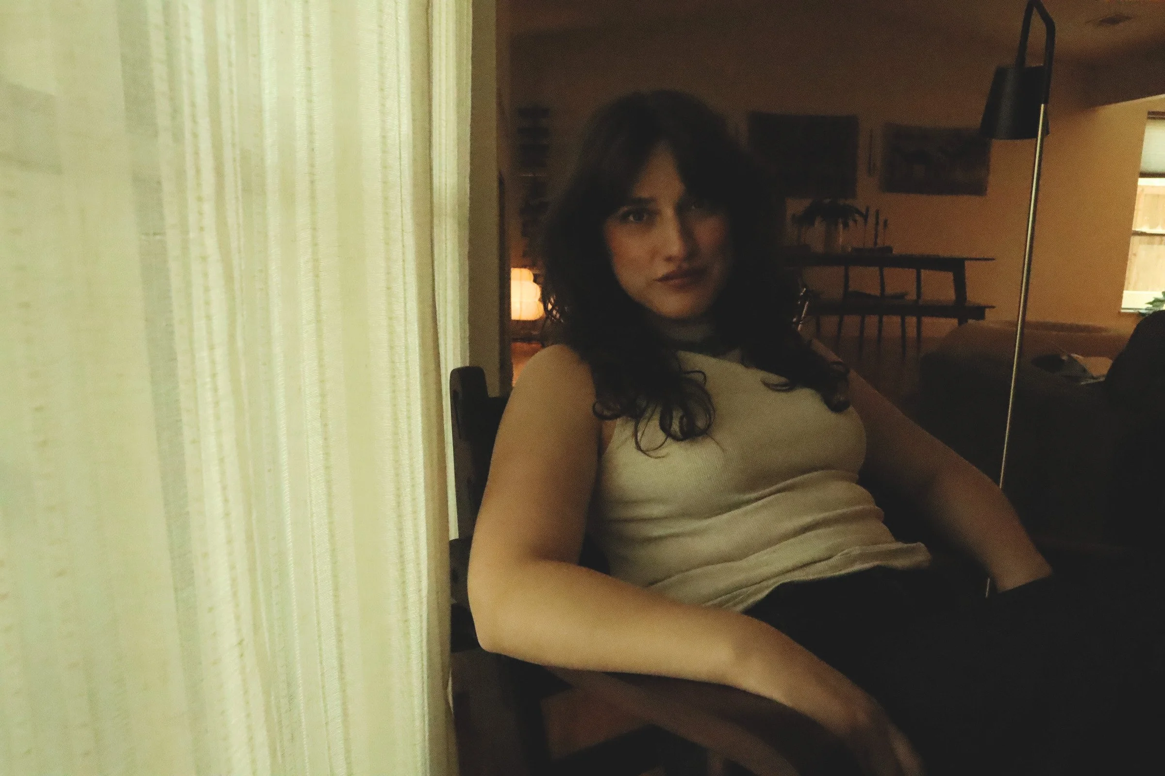A woman with dark, curly hair sitting indoors near a yellow curtain, with a dimly lit background featuring a dining table and decorations.