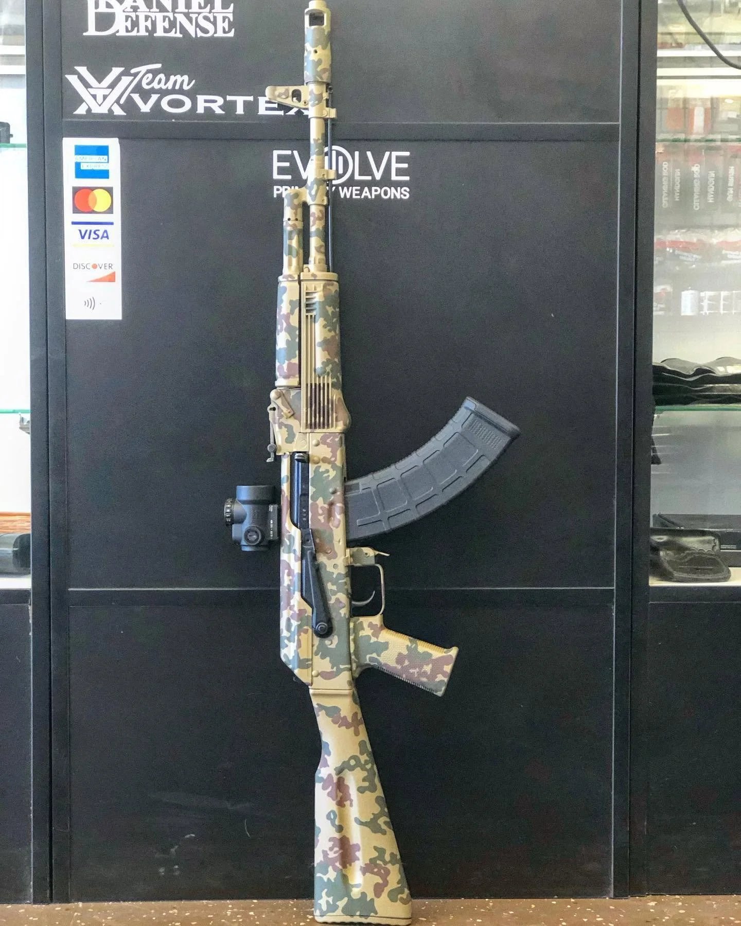 Camouflage-patterned rifle with scope and magazine, standing vertically against a black display case with weapons-related logos and stickers.