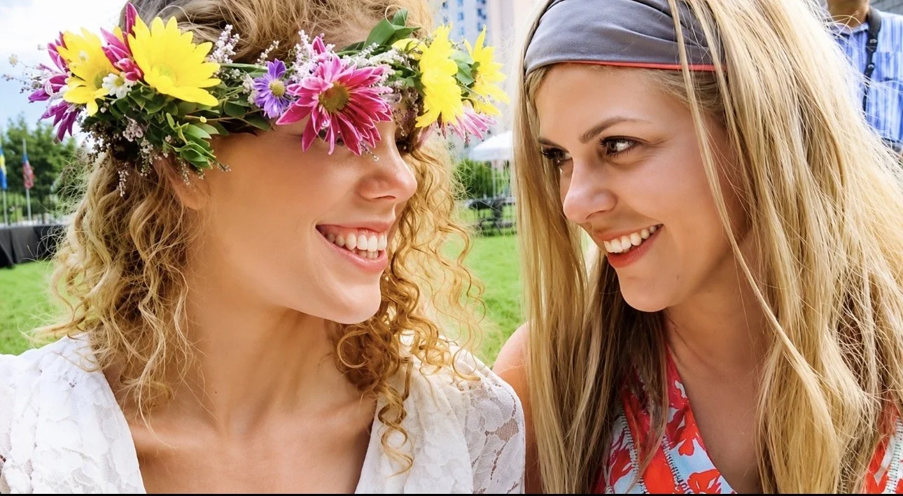 Two women smiling at each other outdoors, one woman wearing a floral crown and a white lace top, the other in a colorful patterned shirt and headband.