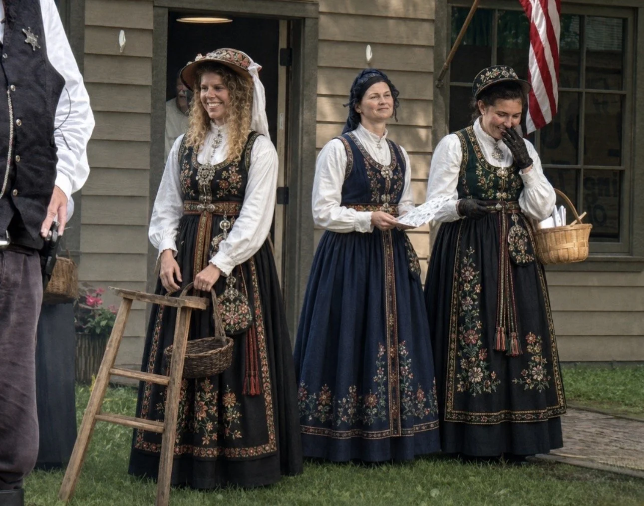 Three women dressed in traditional Norwegian folk costumes standing in front of a wooden building, one woman is holding a piece of paper and another has a wicker basket, and a third wears a bonnet and smiles.