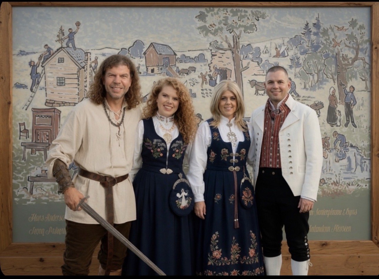Four people in traditional Norwegian clothing standing indoors in front of a large embroidered tapestry depicting a rural scene with houses, trees, people, and animals.