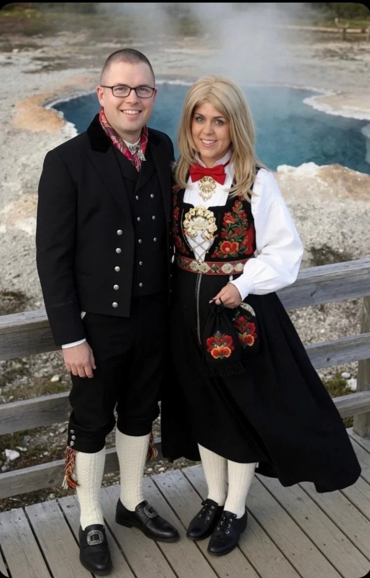 A couple dressed in traditional Norwegian bunad standing on a wooden deck near a geothermal hot spring with steam rising in the background.