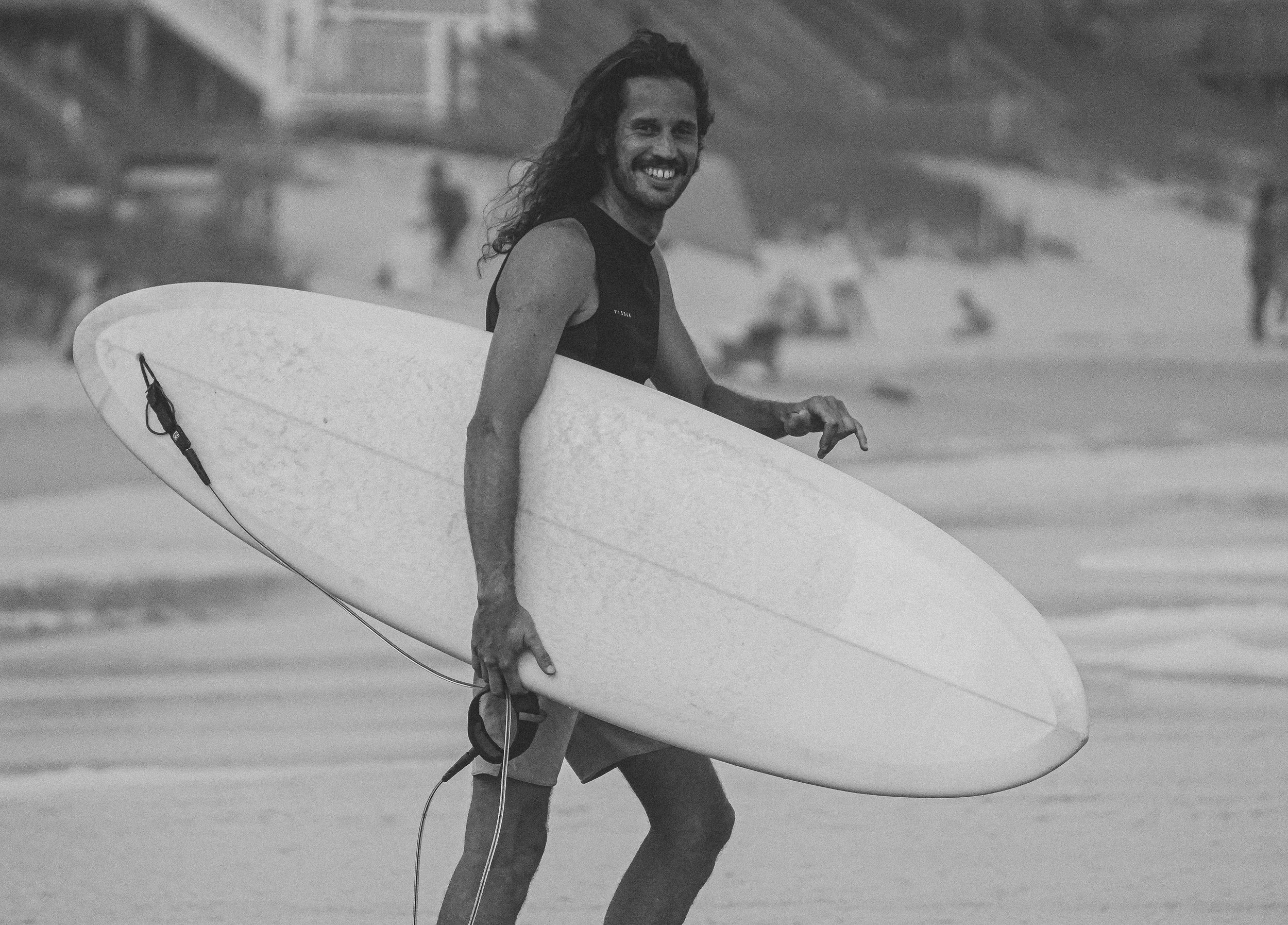 A man with long hair and a beard smiling at the camera, carrying a surfboard while walking on a beach.