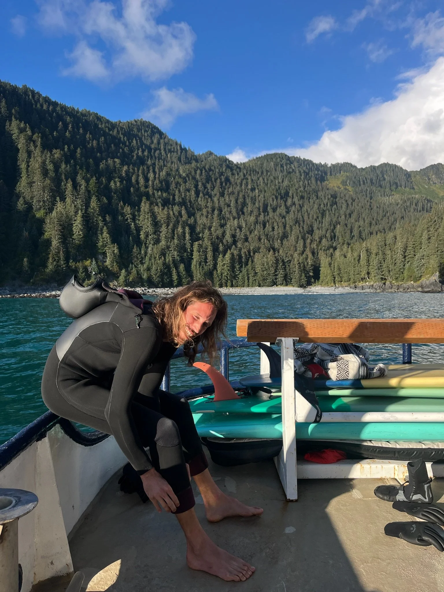 Person bending over on a boat with a mountain landscape in the background, trees on the mountainside, and a wooden bench on the boat with belongings on it.