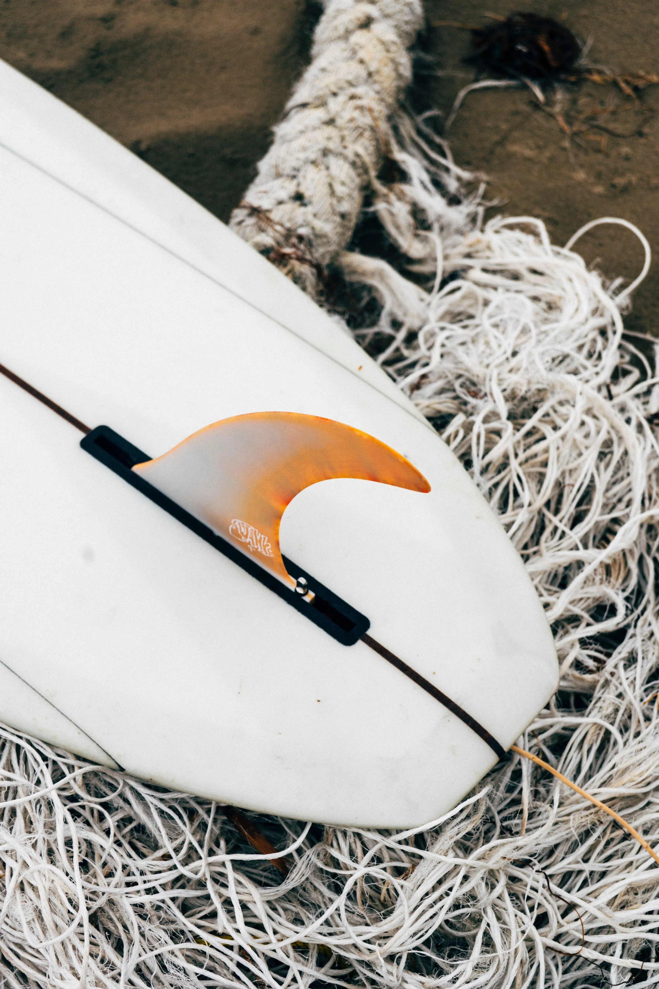A surfboard with a small orange fin resting on top of a pile of tangled white ropes on a sandy surface.