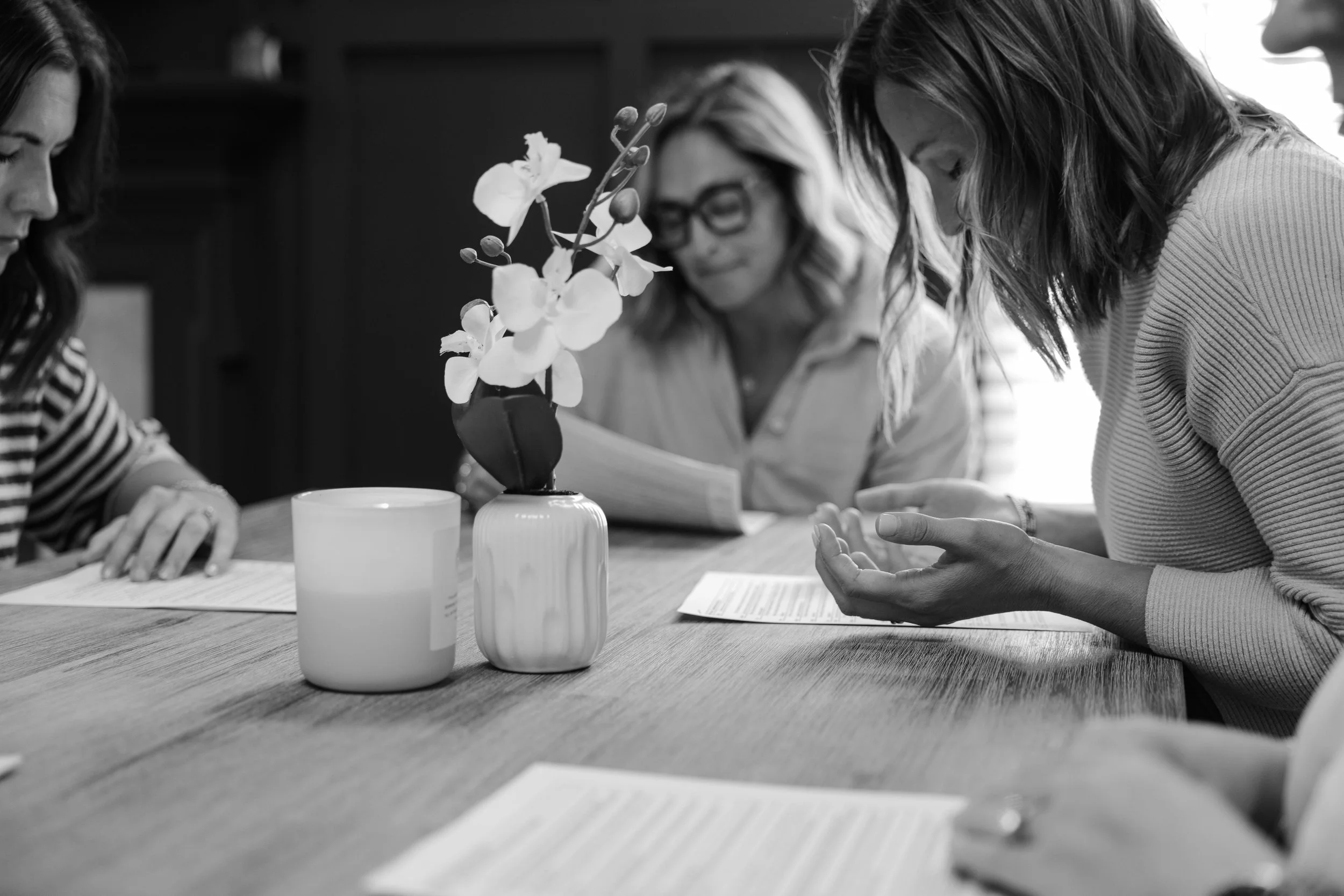 Three women are seated at a table engaged in a discussion, with a vase of flowers and some papers in front of them.