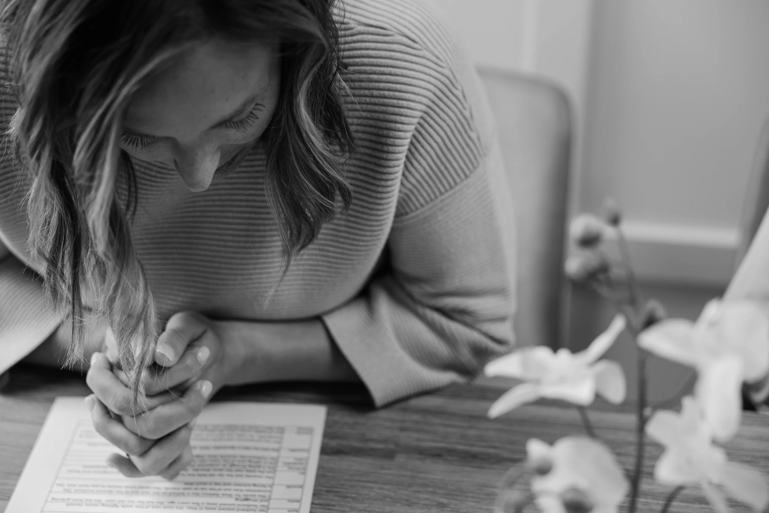 A woman with long hair is filling out paperwork at a table, with a pen in her hand. There are some documents and papers on the table, and the background is blurred with what appears to be a wall or door.